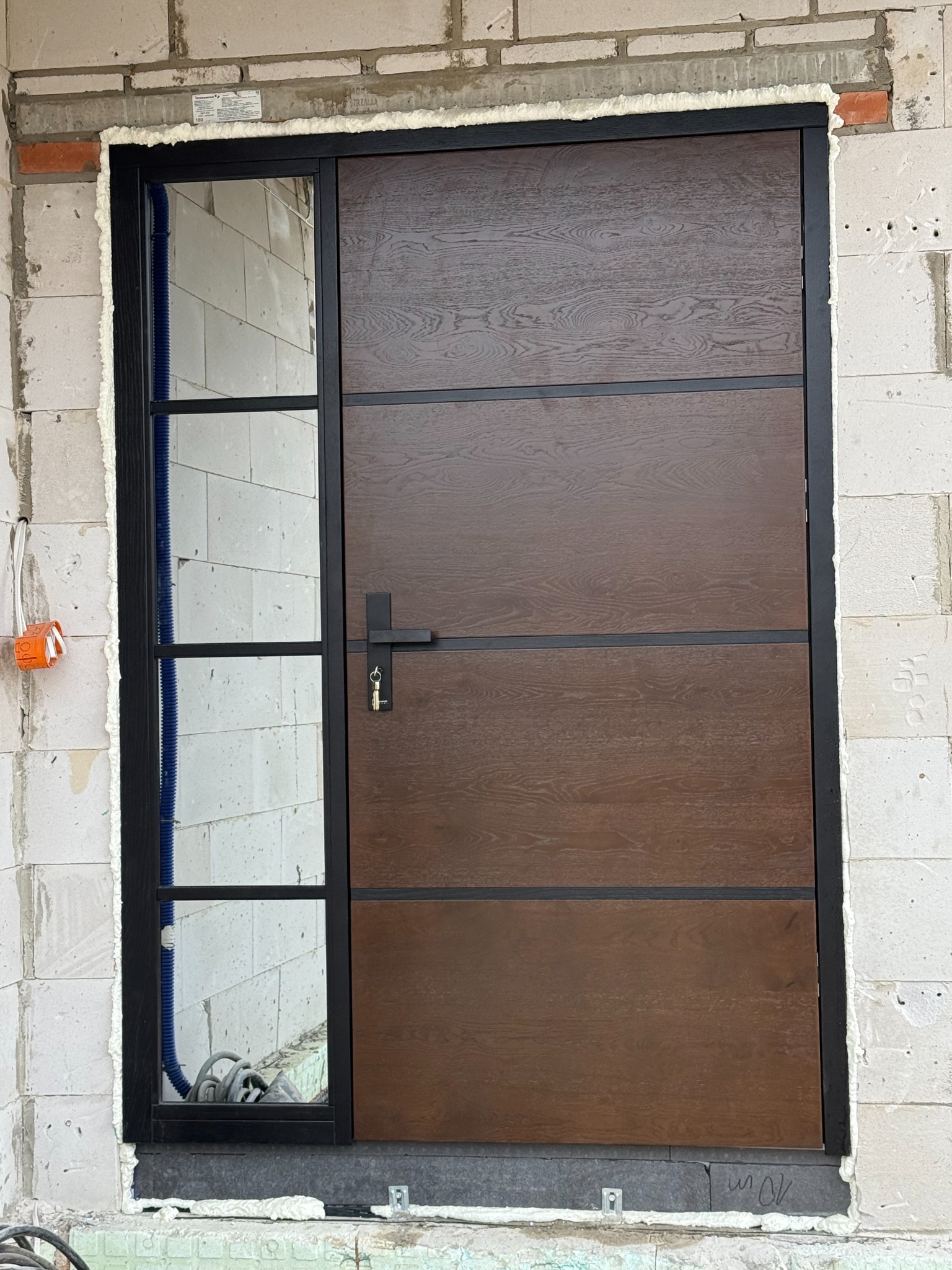 Nowoczesne drzwi zewnętrzne z szybą boczną, brązowe, z czarną ramą, zamontowane w surowym murze w Janowie Lubelskim. Widoczne pianowanie uszczelniające.