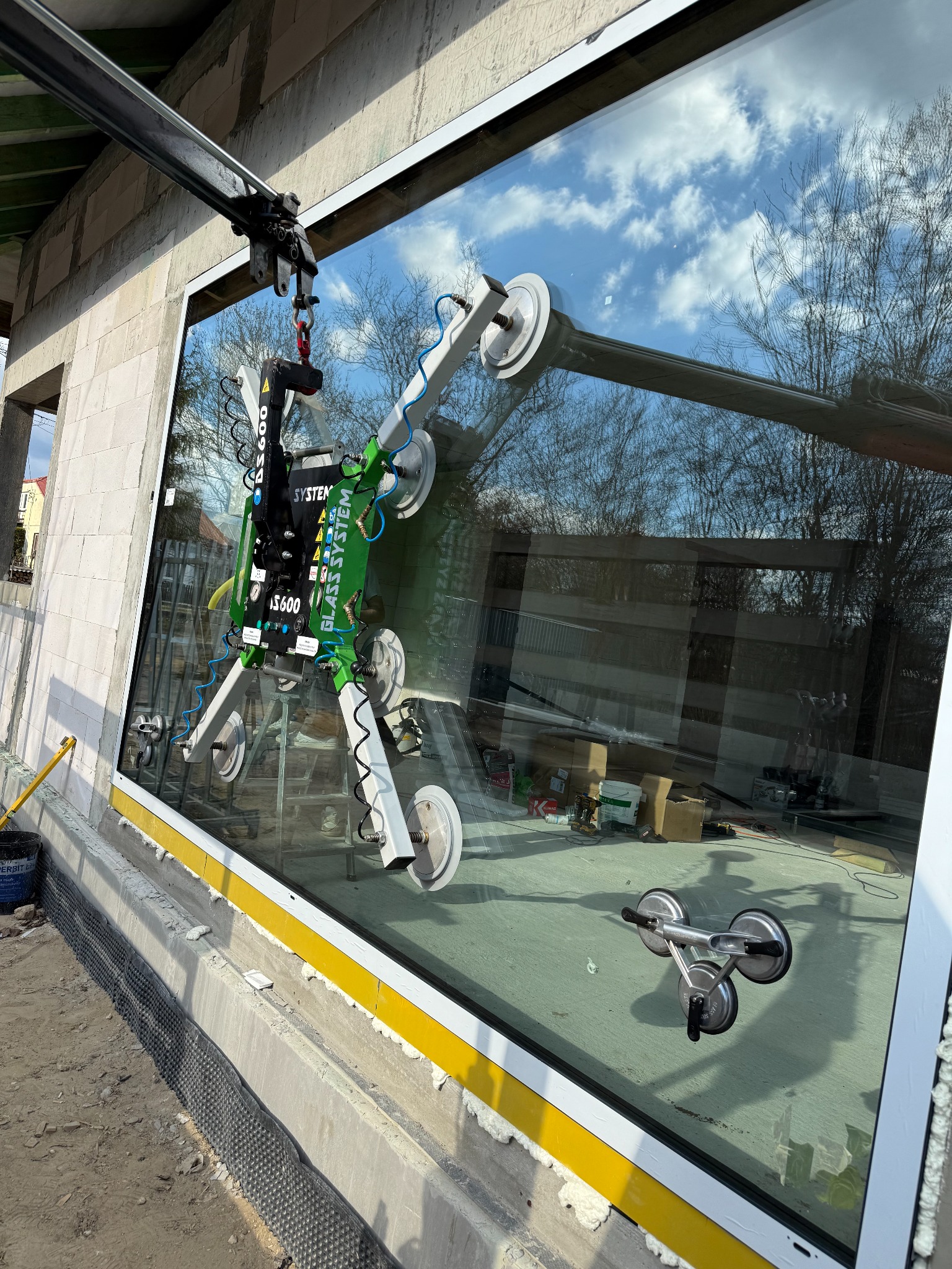 Montaż okna z użyciem przyssawki mechanicznej Glass System DS600 na budowie. W tle widoczne niebo odbijające się w szybie i narzędzia.
