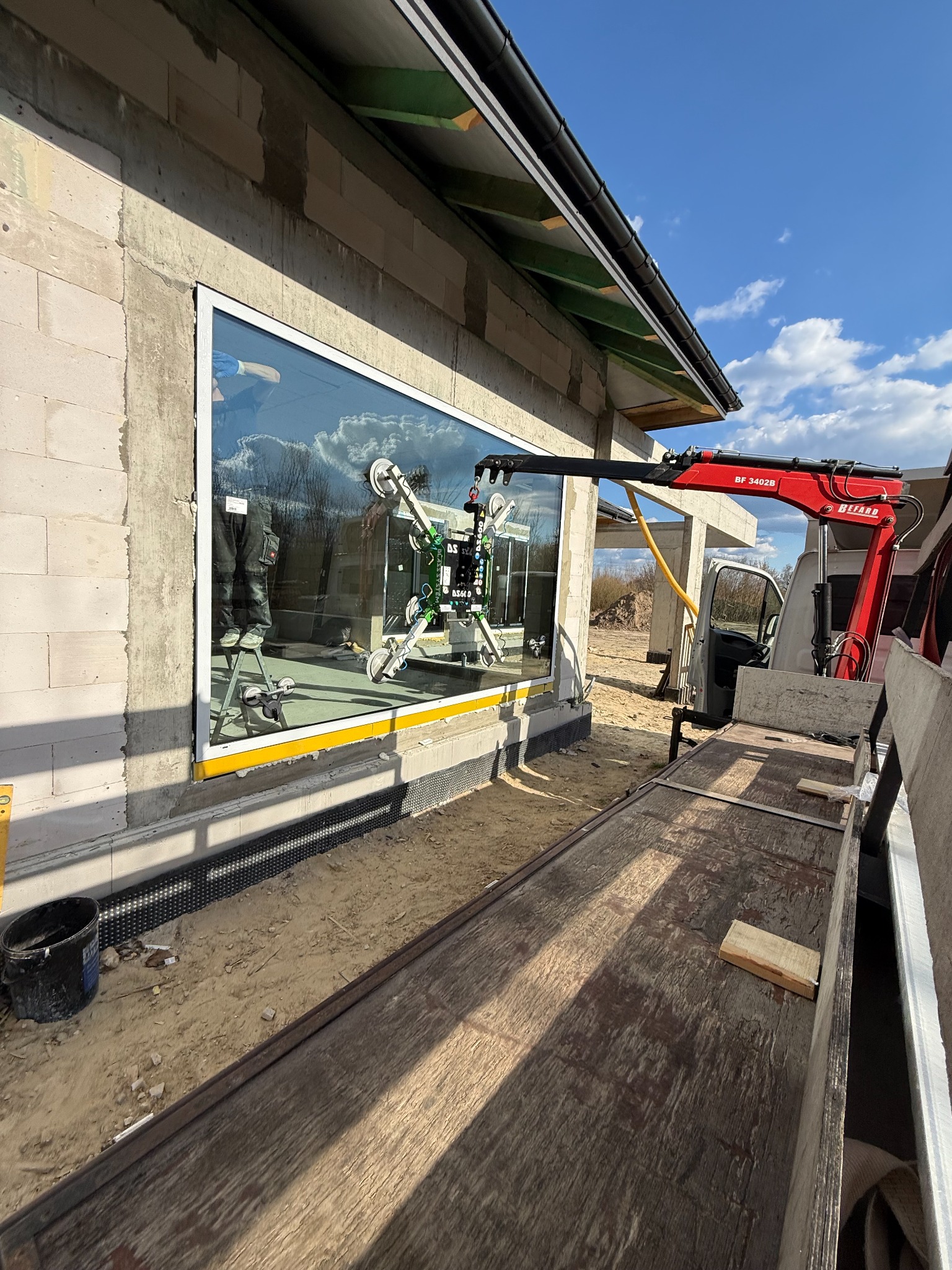 Montaż okna z użyciem przyssawki i żurawia na budowie. Widoczne bloczki betonowe, nieotynkowana ściana i błękitne niebo. Okno duże, z białymi ramami.