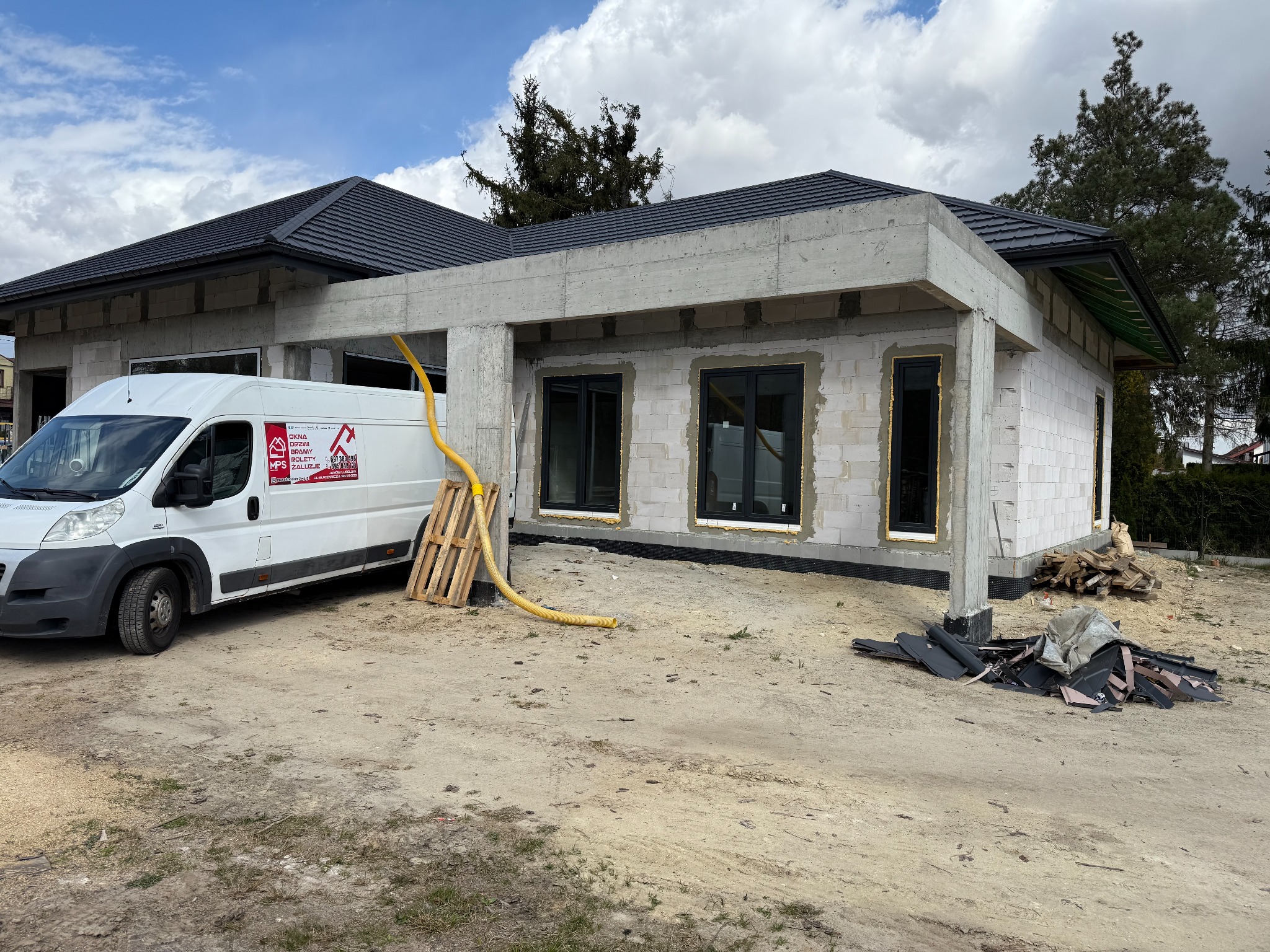 Budynek w stanie surowym z zamontowanymi oknami, na zewnątrz zaparkowany biały samochód dostawczy z logo firmy. Widoczne materiały budowlane i paleta.