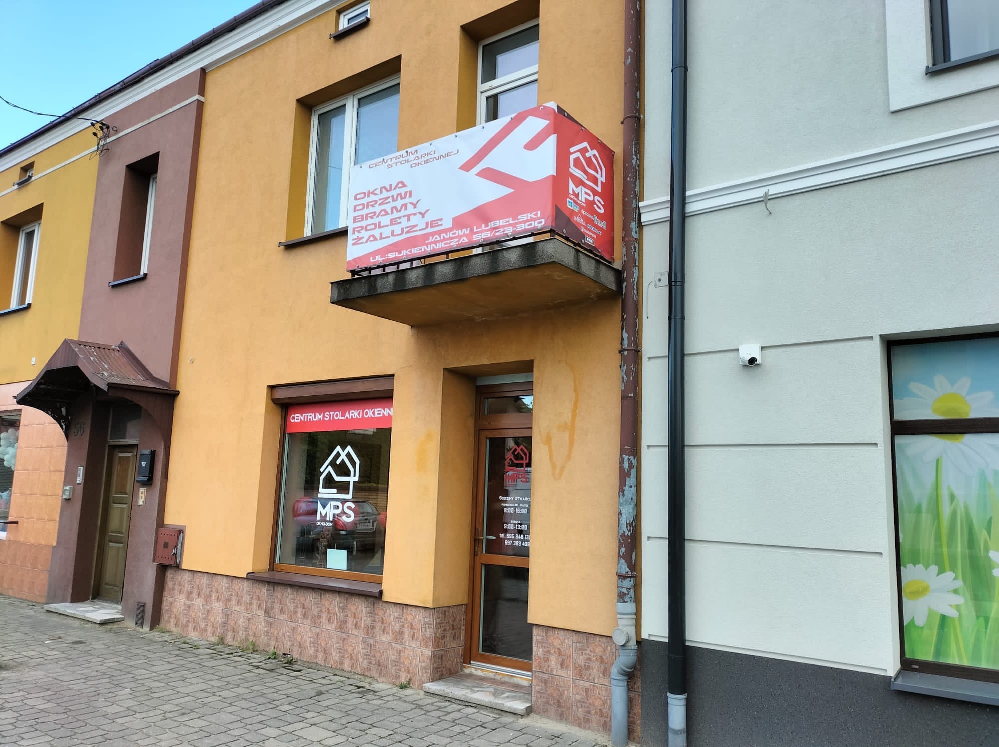 Fasada budynku z reklamą Centrum Stolarki Okiennej MPS z Janowa Lubelskiego, oferującego okna, drzwi, bramy i rolety. Widoczna stolarka okienna i drzwiowa.