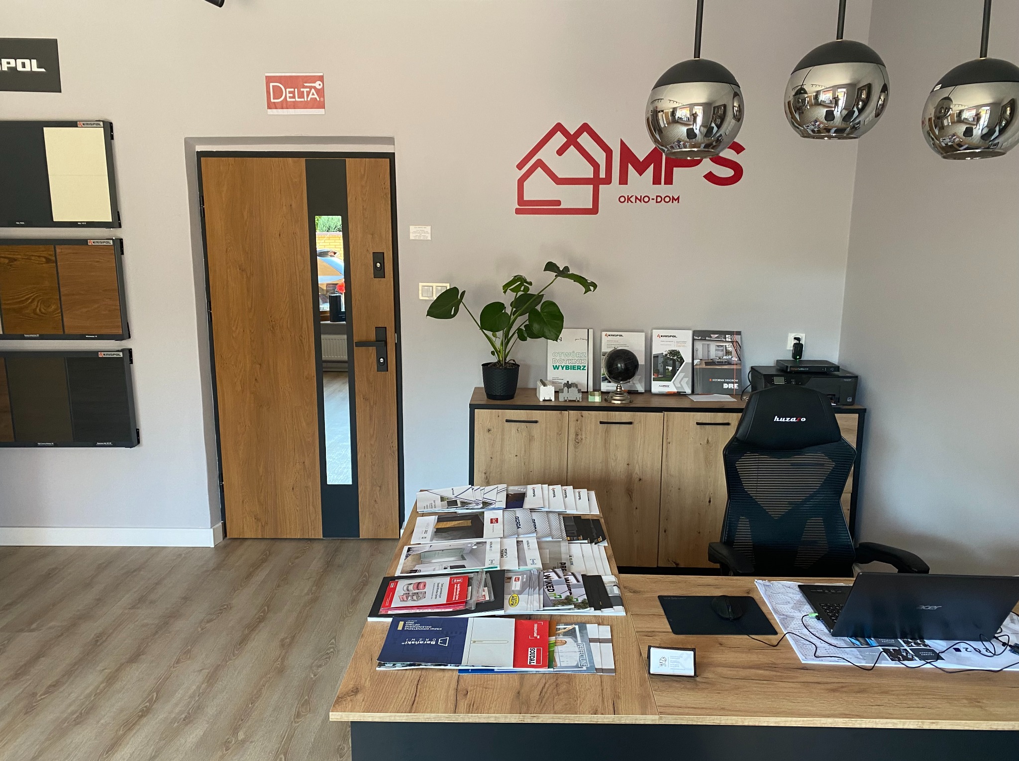 Biuro z ekspozycją drzwi z szybą i logo MPS OKNO-DOM. Na biurku katalogi, laptop i myszka. Wiszące lampy kuliste. W tle próbki materiałów marki Krispol.