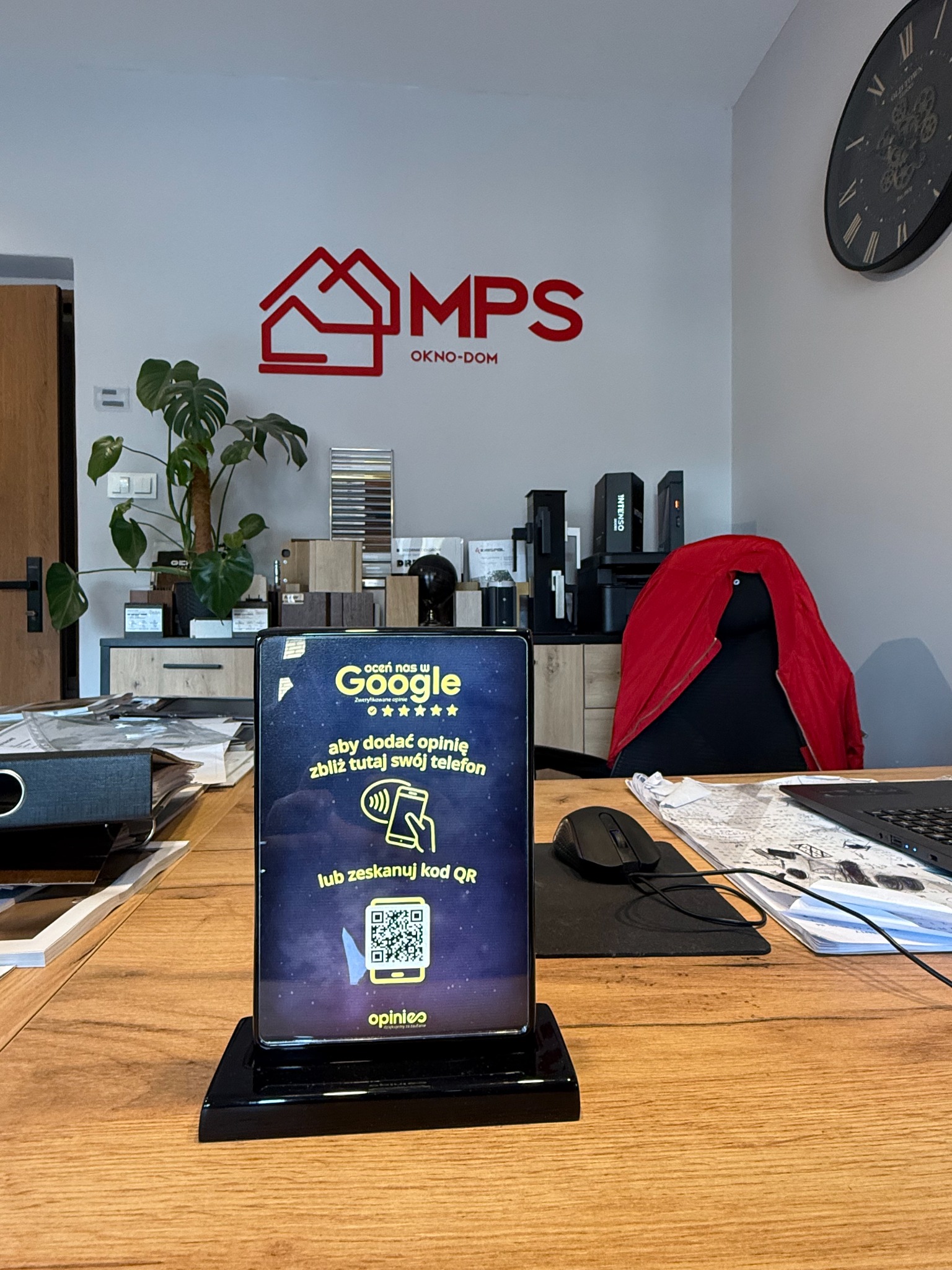Biuro firmy MPS Okno-Dom z logo na ścianie i tabletem z prośbą o ocenę w Google. Na biurku leżą dokumenty i laptop, w tle próbki materiałów.