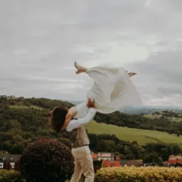 Para młoda w tanecznej pozie na tle malowniczego krajobrazu zalesionych wzgórz i zabudowań. Ujęcie z dołu podkreśla dynamikę i radość chwili.