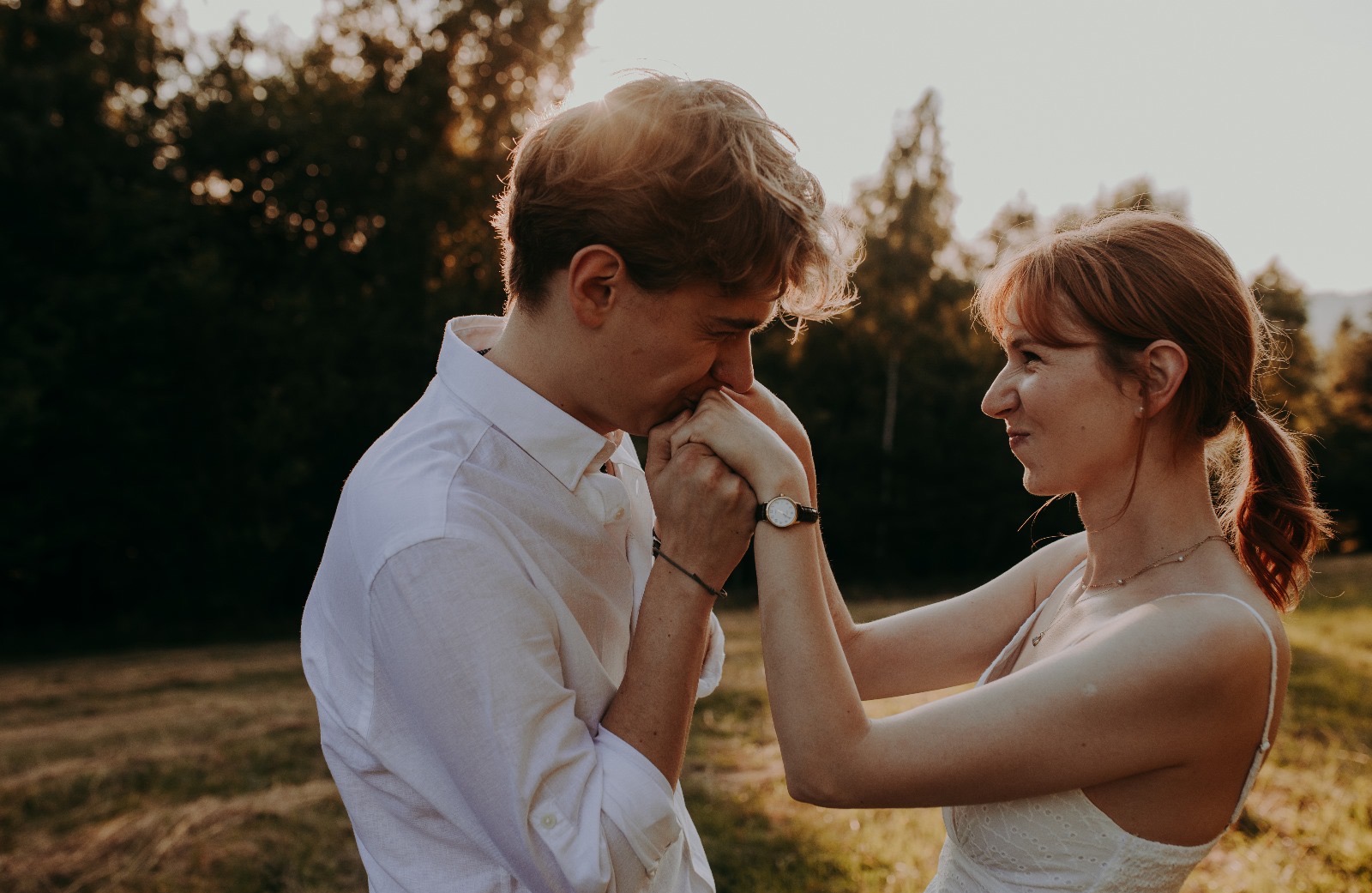 Romantyczna sesja narzeczeńska: Mężczyzna w białej koszuli całuje dłoń uśmiechniętej kobiety z rudymi włosami na tle zieleni i zachodzącego słońca.