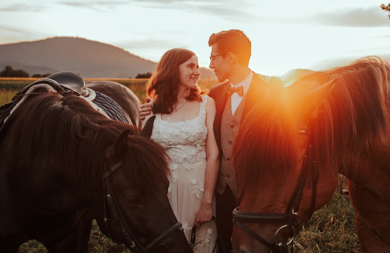 Para młoda w objęciach koni w plenerze, ciepłe światło zachodzącego słońca, wzgórze w tle. Romantyczna sesja ślubna z elementami rustykalnymi.