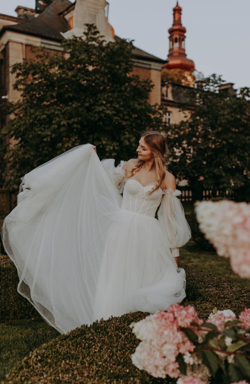 Panna młoda w koronkowej sukni z bufiastymi rękawami, uśmiechnięta, trzyma suknię na tle zieleni i budynku z wieżą. Romantyczna sesja w plenerze.