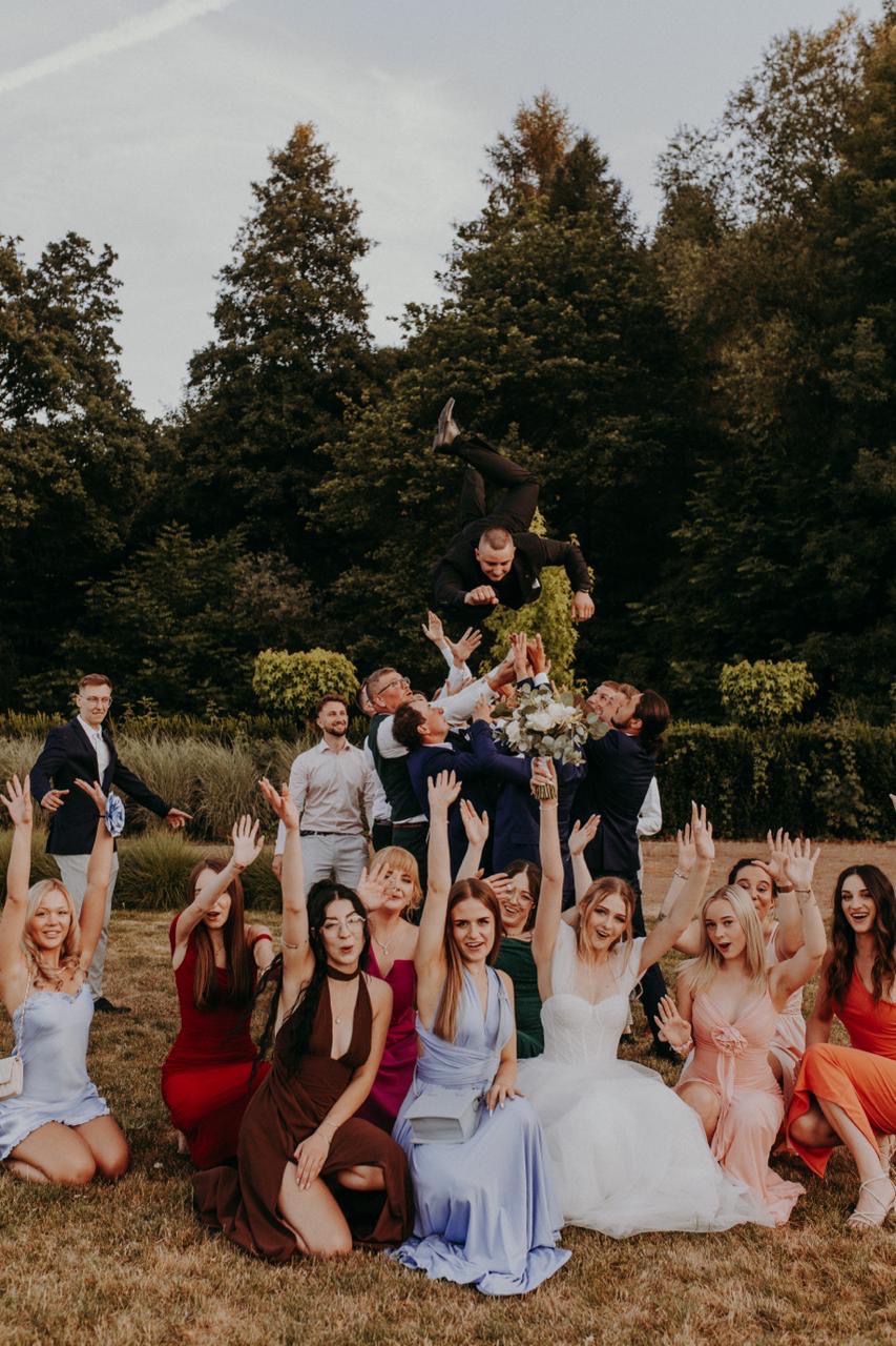 Radosne zdjęcie grupowe z wesela w plenerze: panna młoda w sukni, goście z podniesionymi rękami, jeden z mężczyzn podrzucany w górę na tle zieleni.