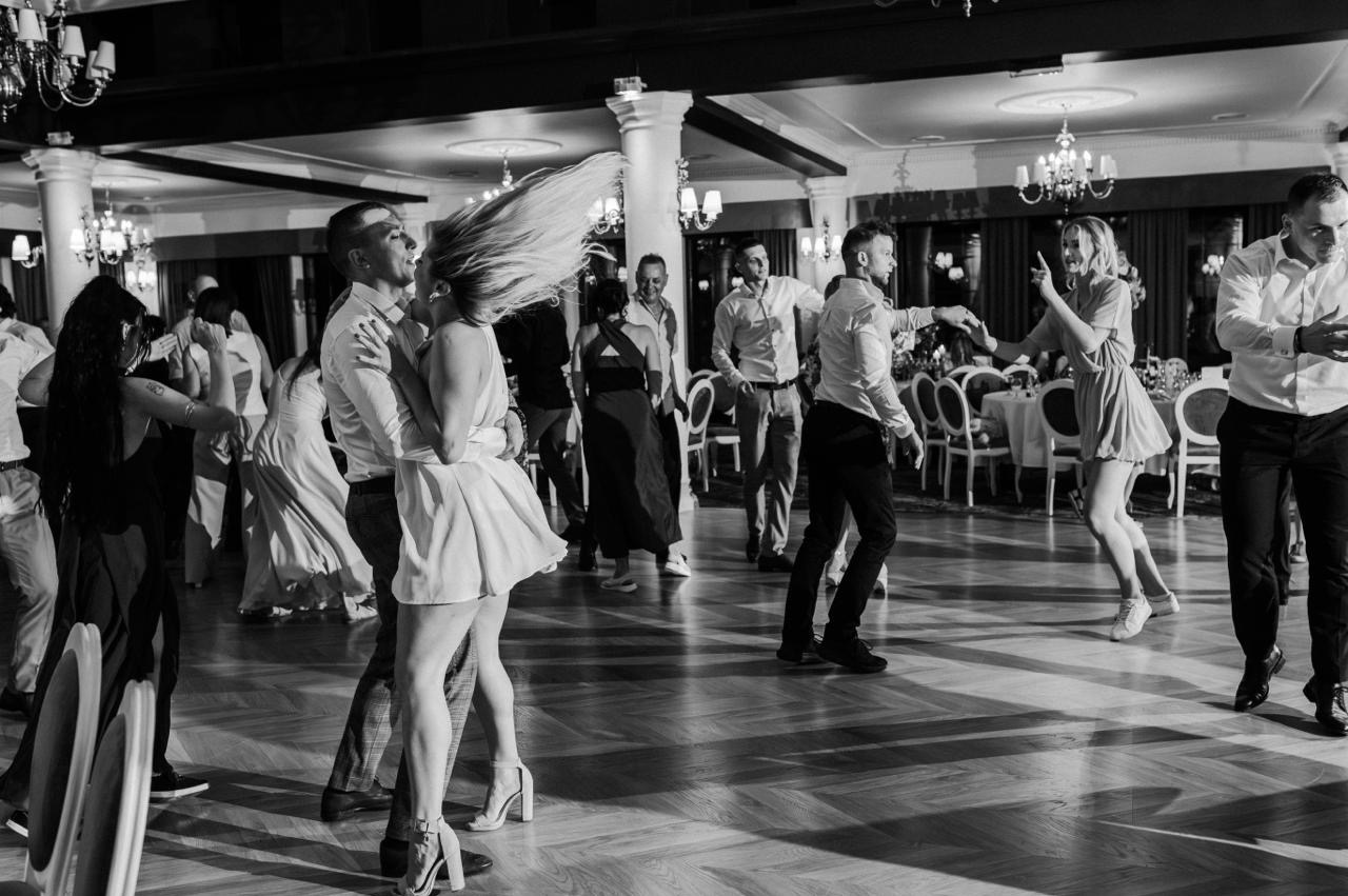 Czarno-białe ujęcie z parkietu sali balowej, gdzie tańczą goście weselni, w tym para w centrum kadru i kobieta z rozrzuconymi włosami.