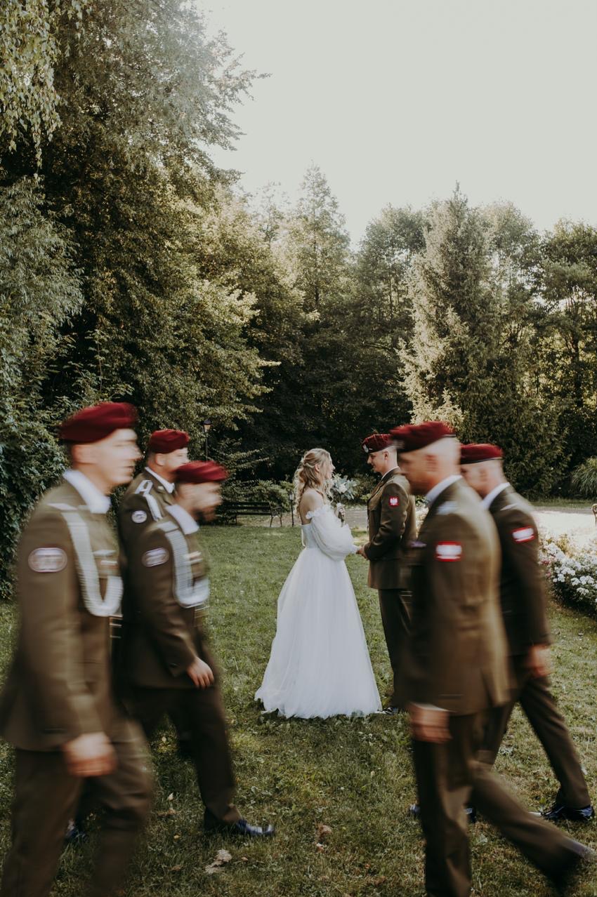 Ślub w plenerze: panna młoda w białej sukni i pan młody w mundurze Wojska Polskiego otoczeni przez żołnierzy w mundurach i czerwonych beretach na tle zieleni.