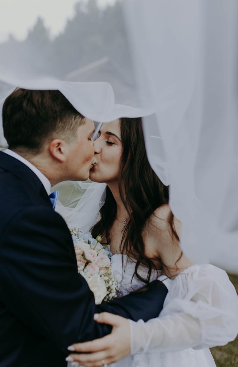 Pocałunek pary młodej pod welonem na tle zieleni. Bukiet w rękach panny młodej, on w granatowym garniturze i błękitną muchą. Ujęcie z bliska, miękkie światło.