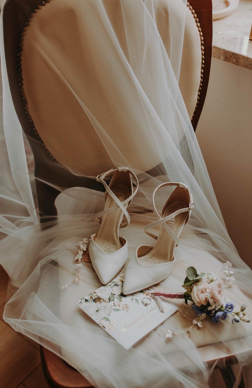 Eleganckie białe szpilki z paskami, zaproszenie ślubne, obrączka i bukiecik na krześle, przykryte delikatnym woalem. Subtelne detale ślubne w ciepłej tonacji.