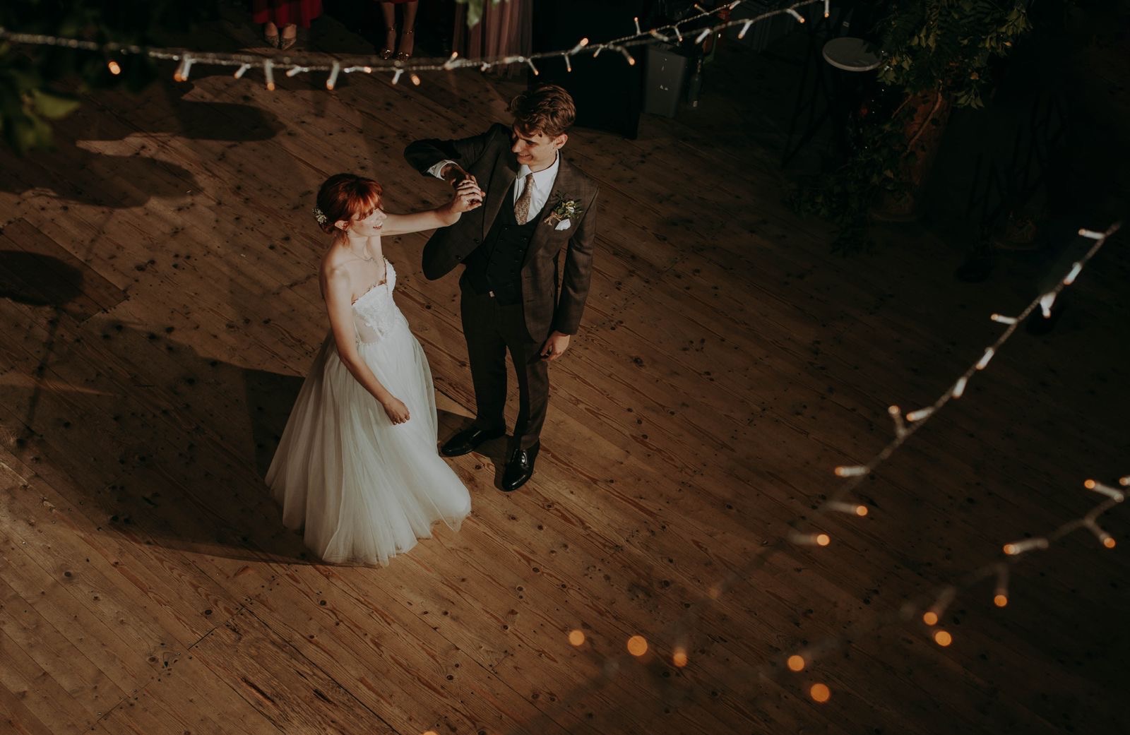 Para młoda w tańcu na parkiecie z drewna, oświetlonym girlandami. Panna młoda w białej sukni, pan młody w brązowym garniturze. Ujęcie z góry, ciepłe światło.