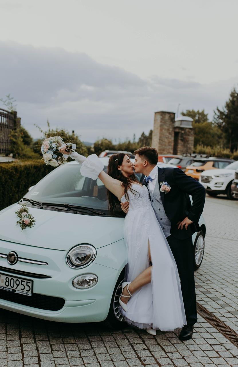 Uśmiechnięta para młoda całuje się przy turkusowym Fiacie 500 z dekoracjami kwiatowymi, na tle parkingu i ceglanej wieży. Bukiet w ręku panny młodej.