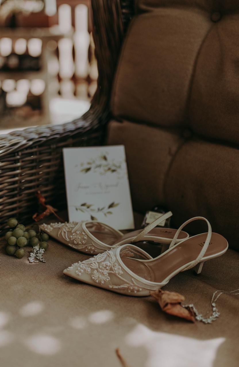 Eleganckie, zdobione perłami buty ślubne na brązowym tle, obok zaproszenie, kolczyki i naszyjnik. Ujęcie z bliska, ciepłe światło.