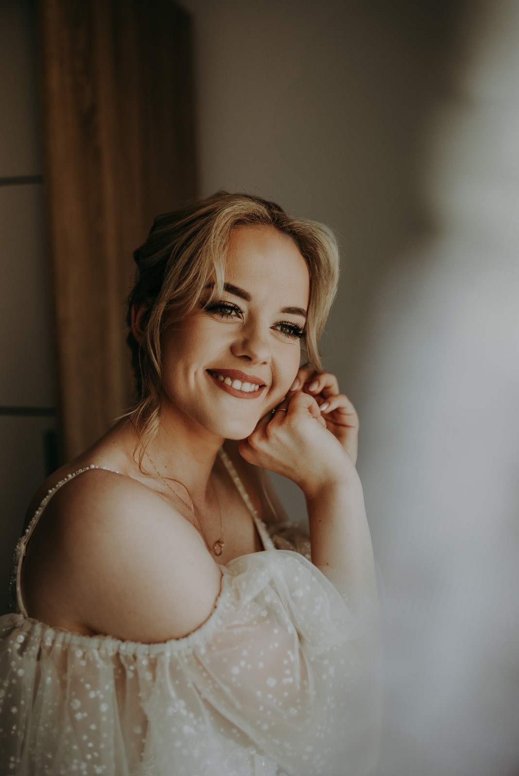 Uśmiechnięta blondynka w sukni z delikatnej siateczki, poprawia kolczyk. Portret w ciepłych tonach, z naturalnym światłem padającym z boku.