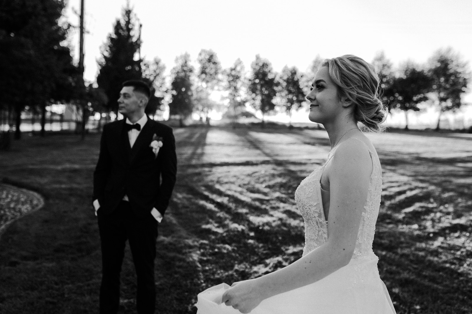 Czarno-białe zdjęcie pary młodej w plenerze: panna młoda z profilu w koronkowej sukni, pan młody w tle w eleganckim smokingu, drzewa i trawa w tle.