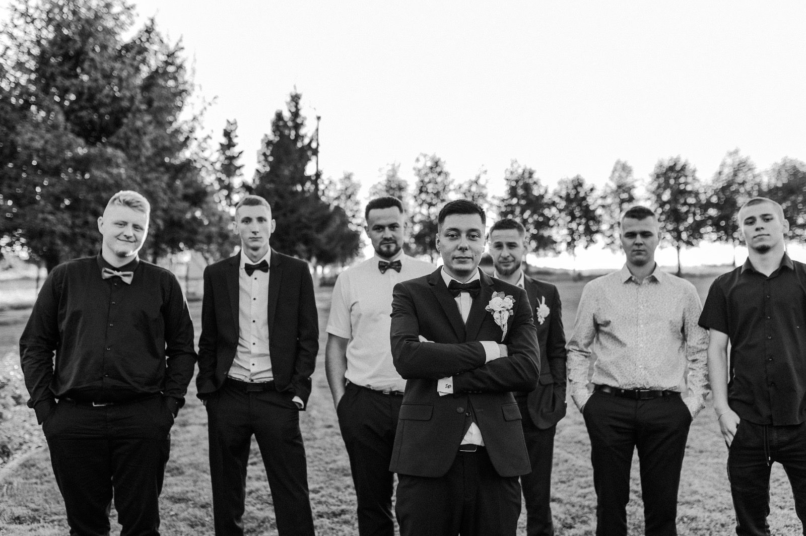 Czarno-białe zdjęcie grupy mężczyzn w eleganckich strojach, z których jeden wyróżnia się na pierwszym planie z założonymi rękami, na tle drzew i otwartej przestrzeni.