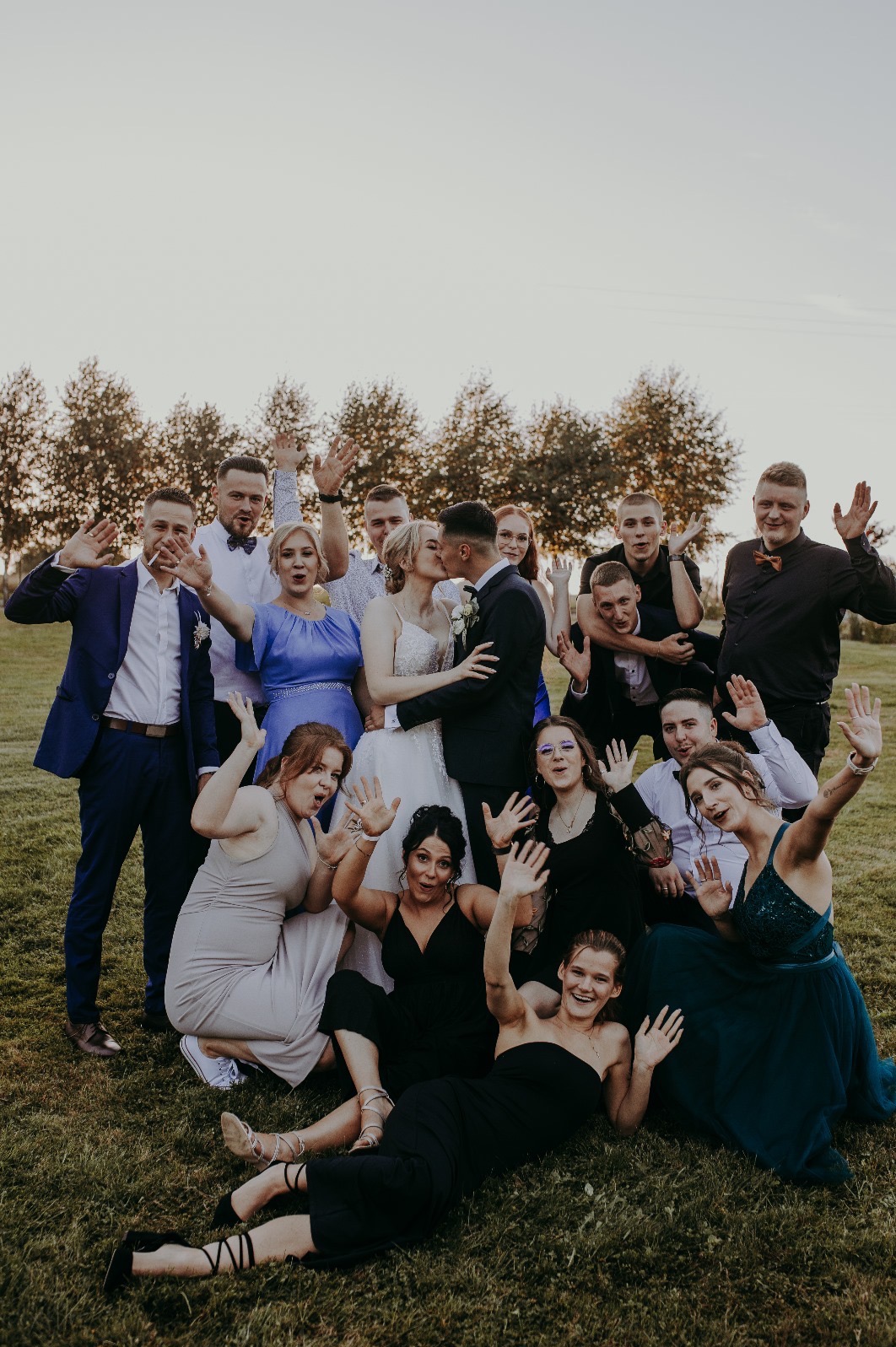 Para młoda całuje się otoczona gośćmi weselnymi na zielonej trawie w Żywcu. Wszyscy machają radośnie do obiektywu, drzewa w tle.