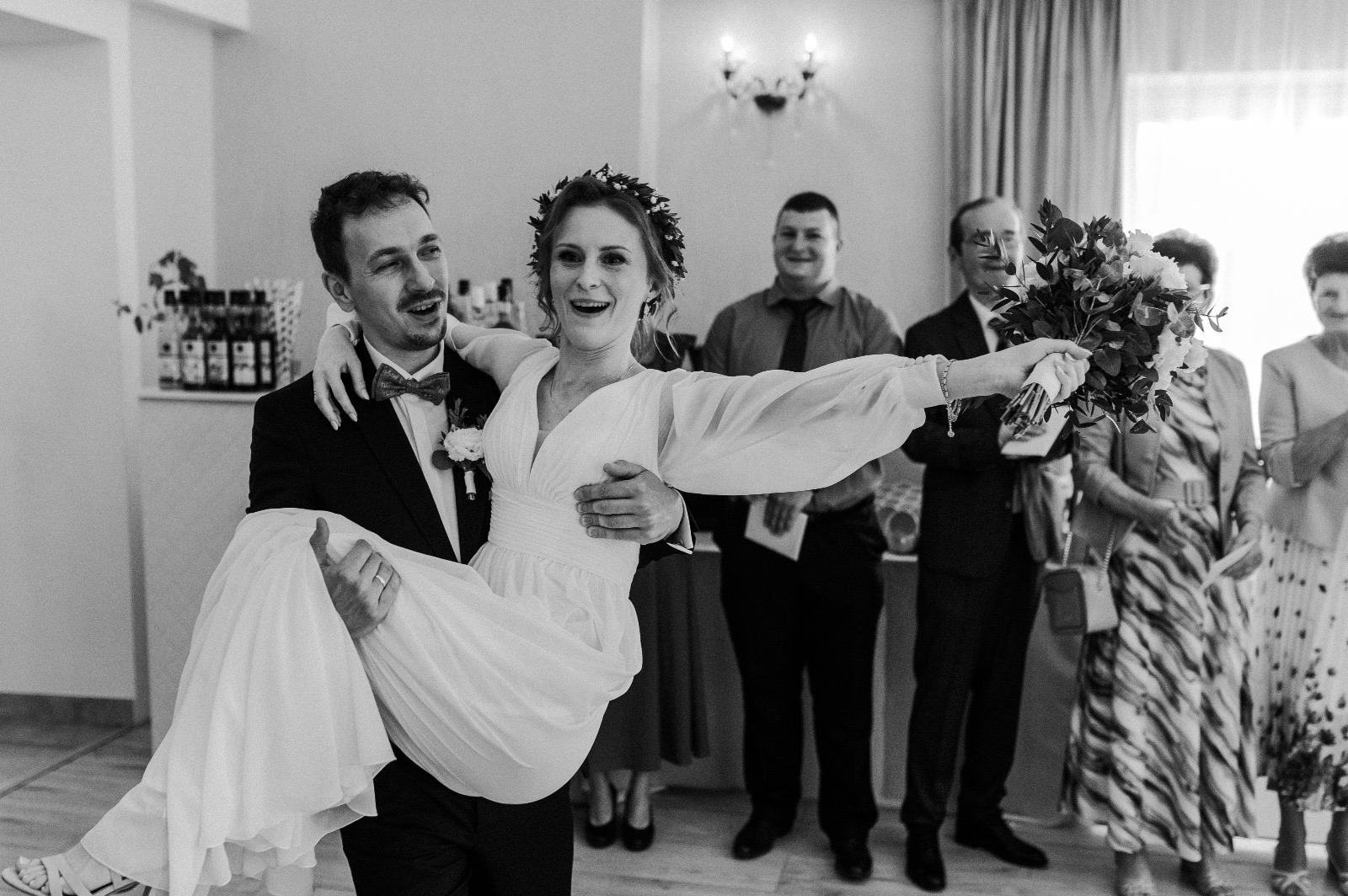 Czarno-białe zdjęcie: Pan młody trzyma na rękach uśmiechniętą pannę młodą w sukni z bukietem. W tle goście weselni.