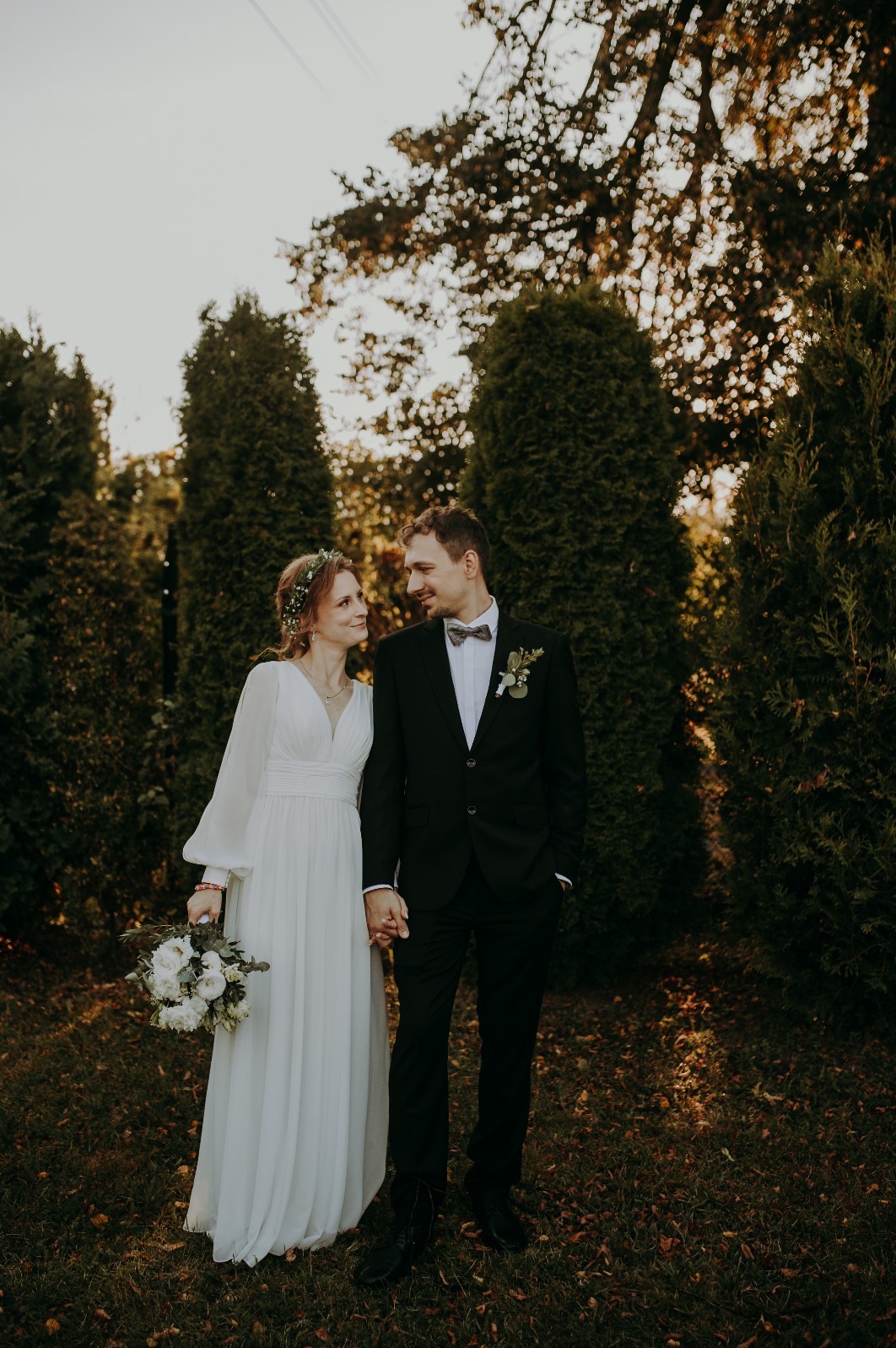 Para młoda trzymająca się za ręce, w tle zielone tuje i zachodzące słońce. Ona w zwiewnej sukni, on w czarnym garniturze z muchą. Subtelne oświetlenie.