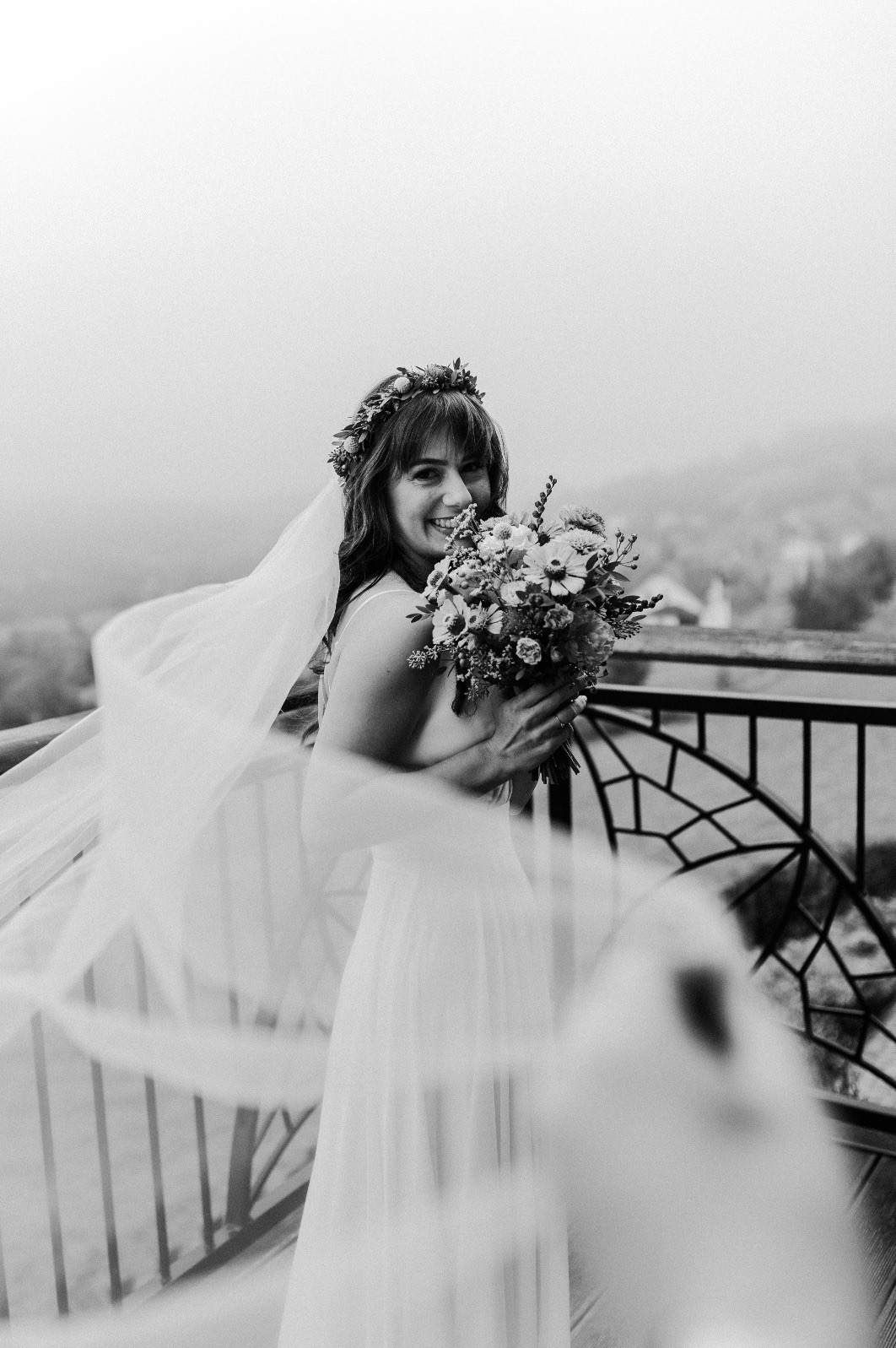 Uśmiechnięta panna młoda w koronie z kwiatów i z bukietem, na tle metalowej balustrady i z welonem na wietrze, czarno-białe zdjęcie.