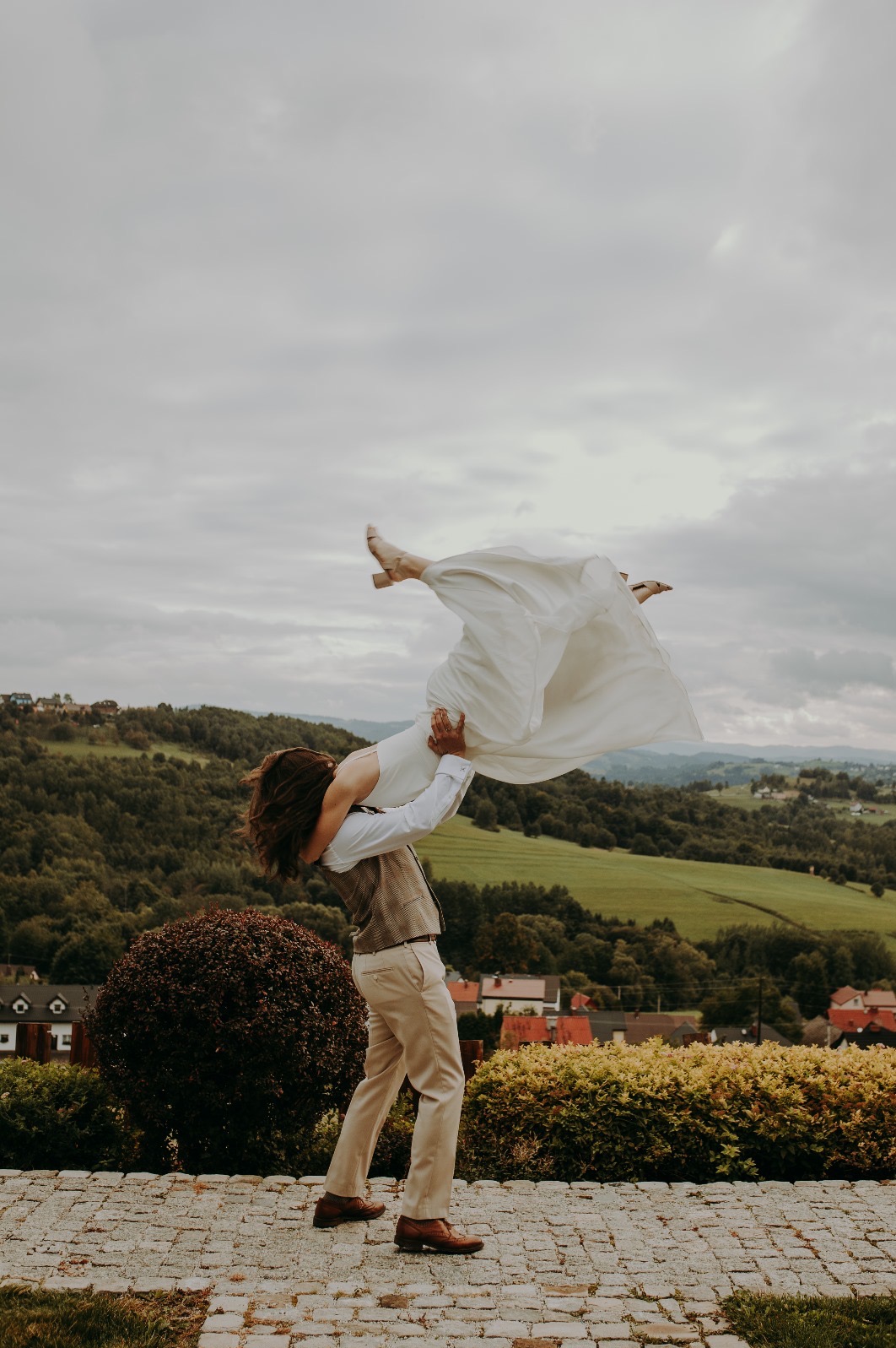 Para młoda w tanecznej pozie na tle malowniczego krajobrazu zalesionych wzgórz i zabudowań. Ujęcie z dołu podkreśla dynamikę i radość chwili.