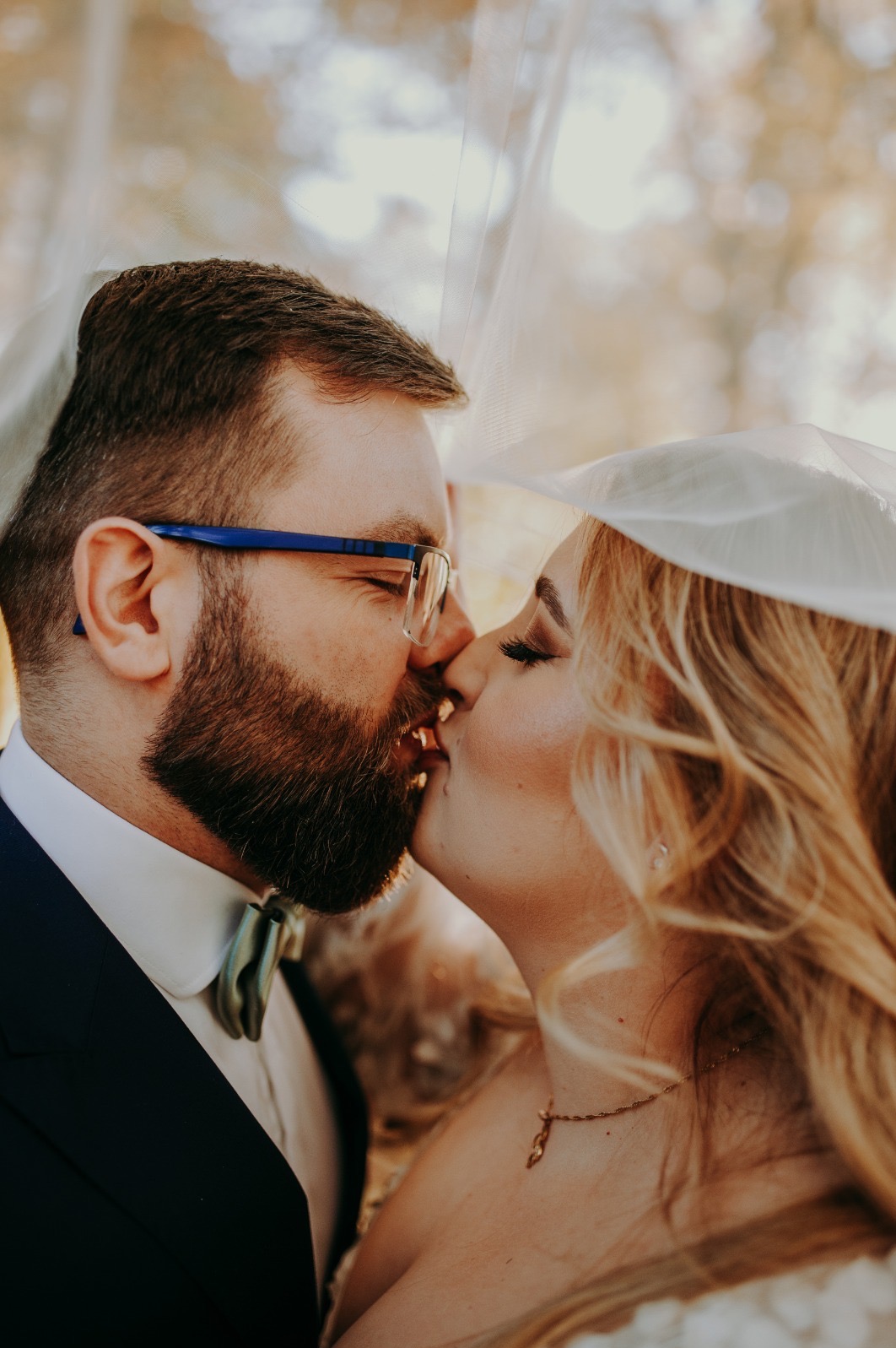 Delikatne zbliżenie na całującą się parę młodą pod welonem. Pan młody w okularach z niebieskimi oprawkami i muszką. Romantyczna atmosfera, ciepłe światło.