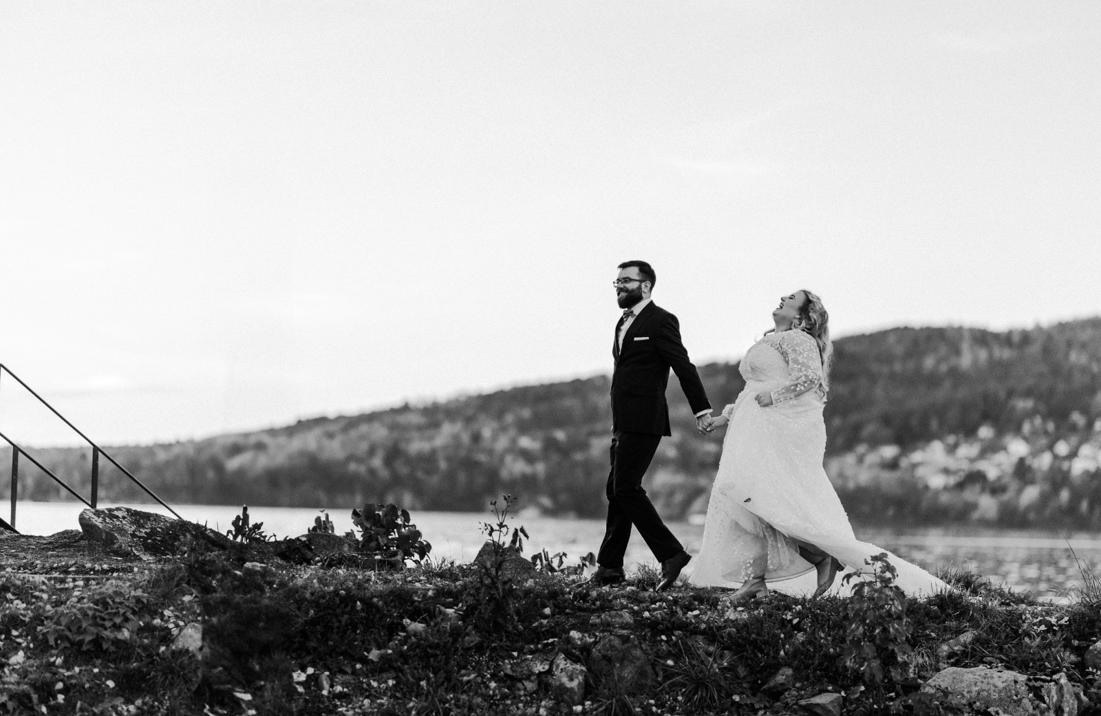 Czarno-białe zdjęcie pary młodej spacerującej brzegiem jeziora. Pan młody w garniturze, panna młoda w sukni z koronkowymi rękawami, oboje uśmiechnięci, trzymają się za ręce.