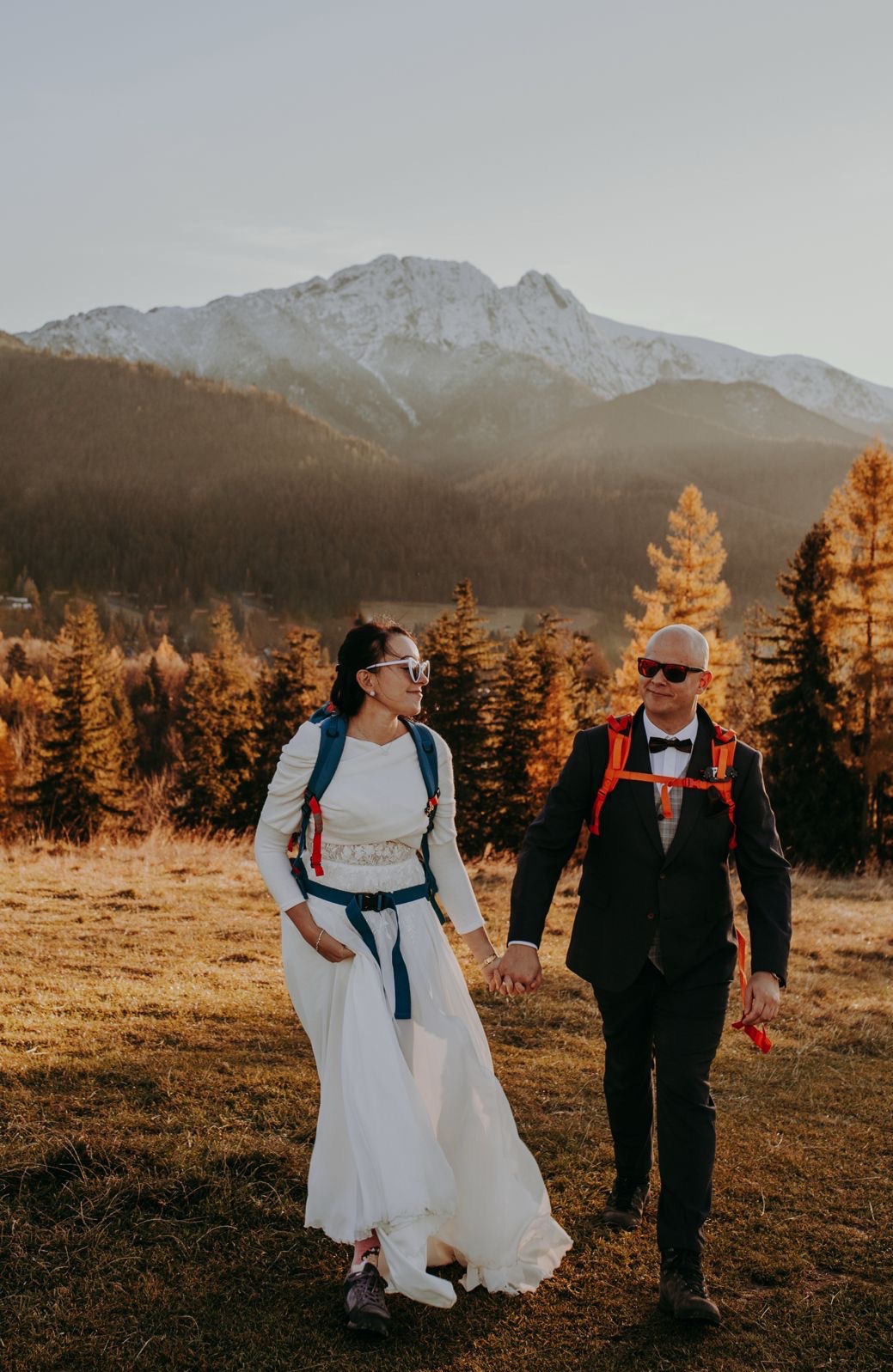 Para młoda w strojach ślubnych i z plecakami trekkingowymi, trzymająca się za ręce na tle górskiego krajobrazu w ciepłym, zachodzącym słońcu. Nietypowa sesja ślubna.