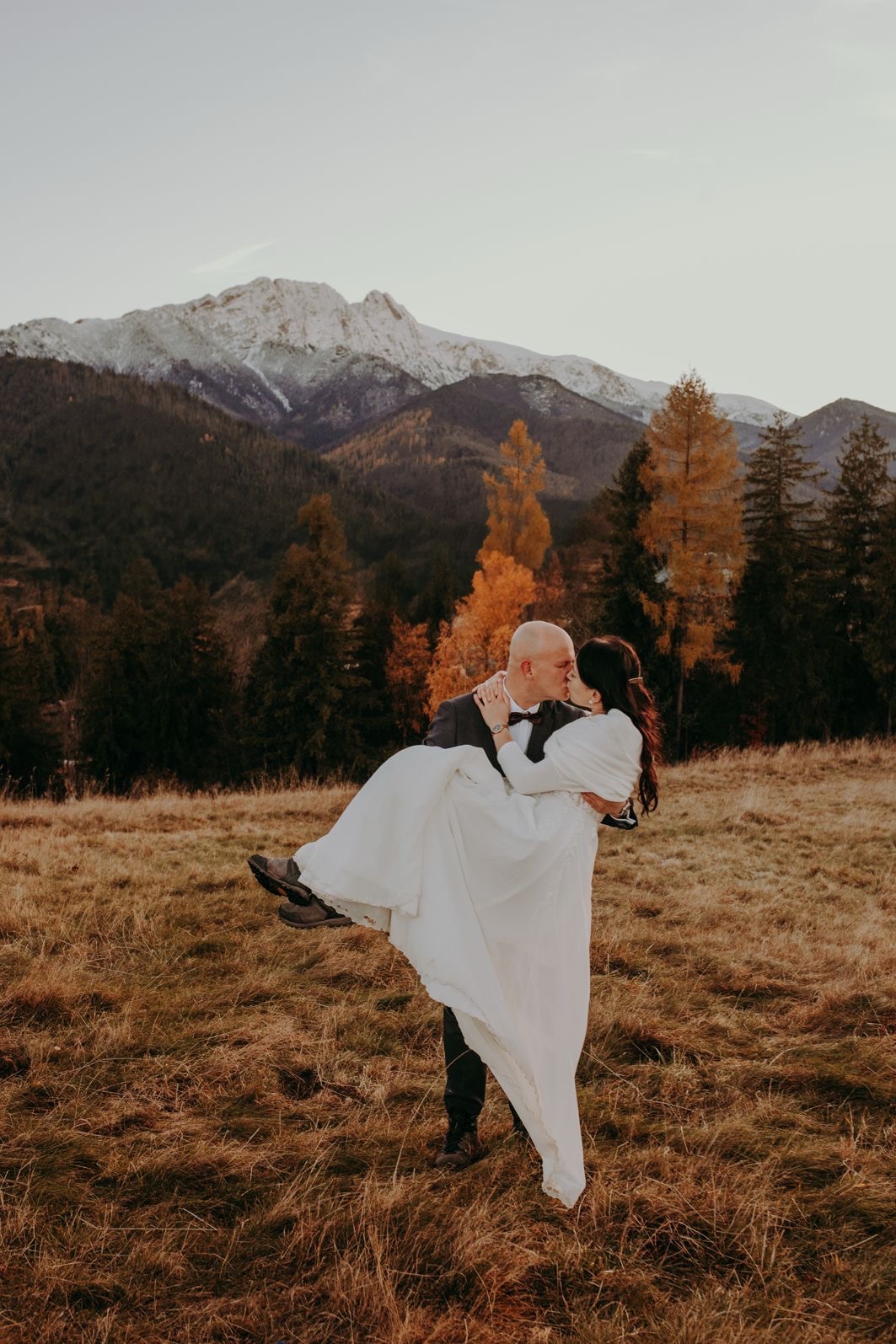 Para młoda w górach: Pan młody trzyma pannę młodą na rękach, całując się na tle górskiego krajobrazu z lasami i ośnieżonym szczytem. Ciepłe, naturalne światło.