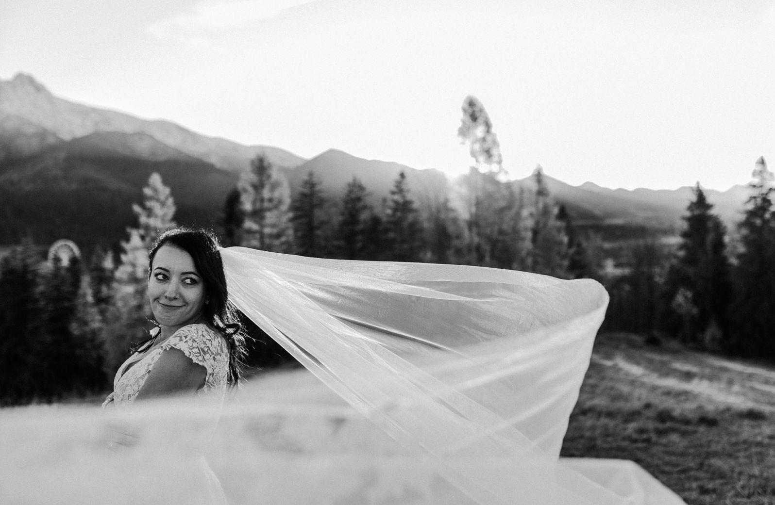 Czarno-białe zdjęcie panny młodej w zwiewnym welonie na tle gór i lasu, z uśmiechem spoglądającej w bok. Delikatne światło podkreśla naturalność scenerii.