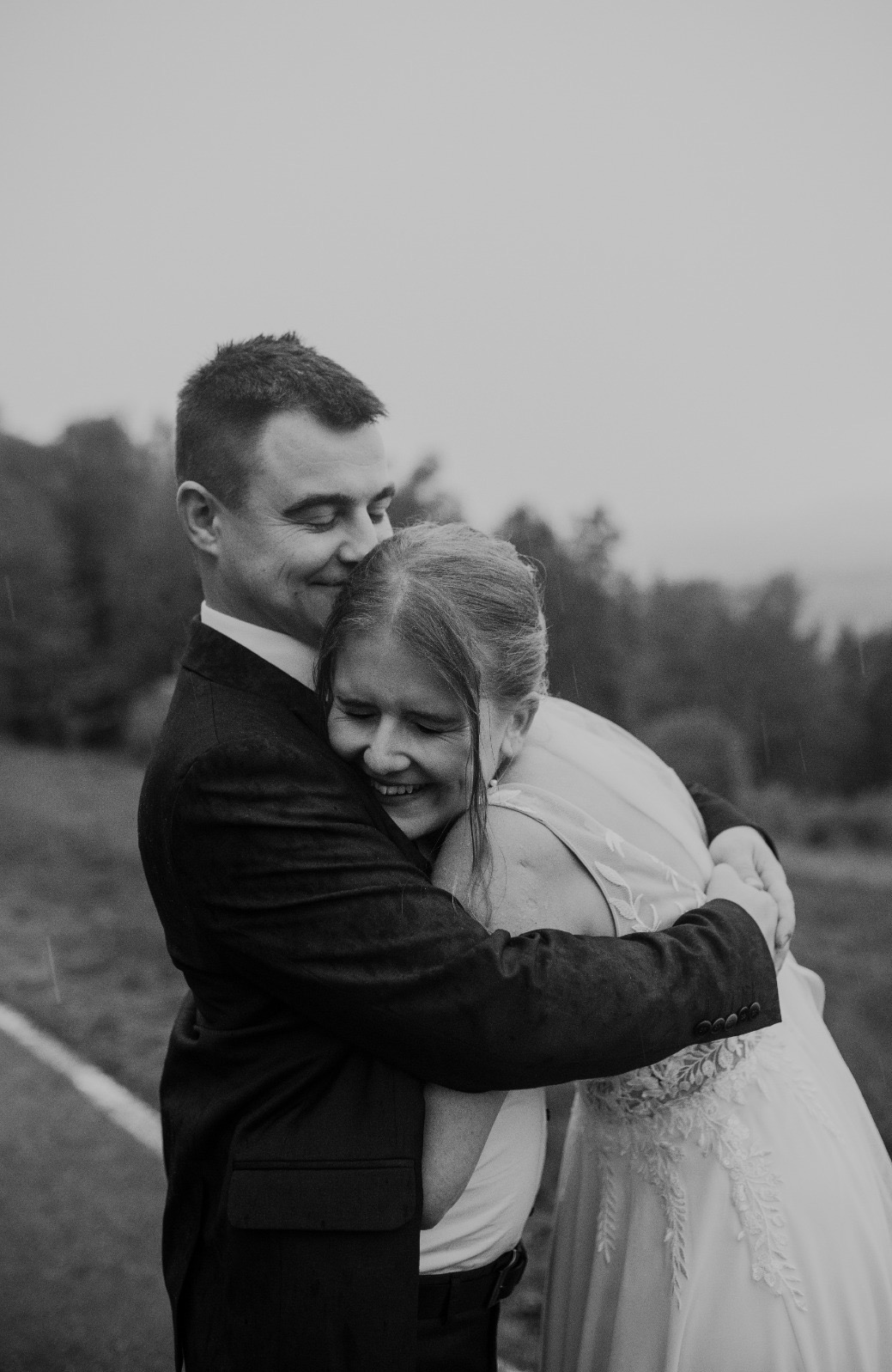 Czarno-białe zdjęcie uśmiechniętej pary młodej w objęciach na tle zalesionego wzgórza w deszczowy dzień. Suknia panny młodej z koronkowymi detalami.