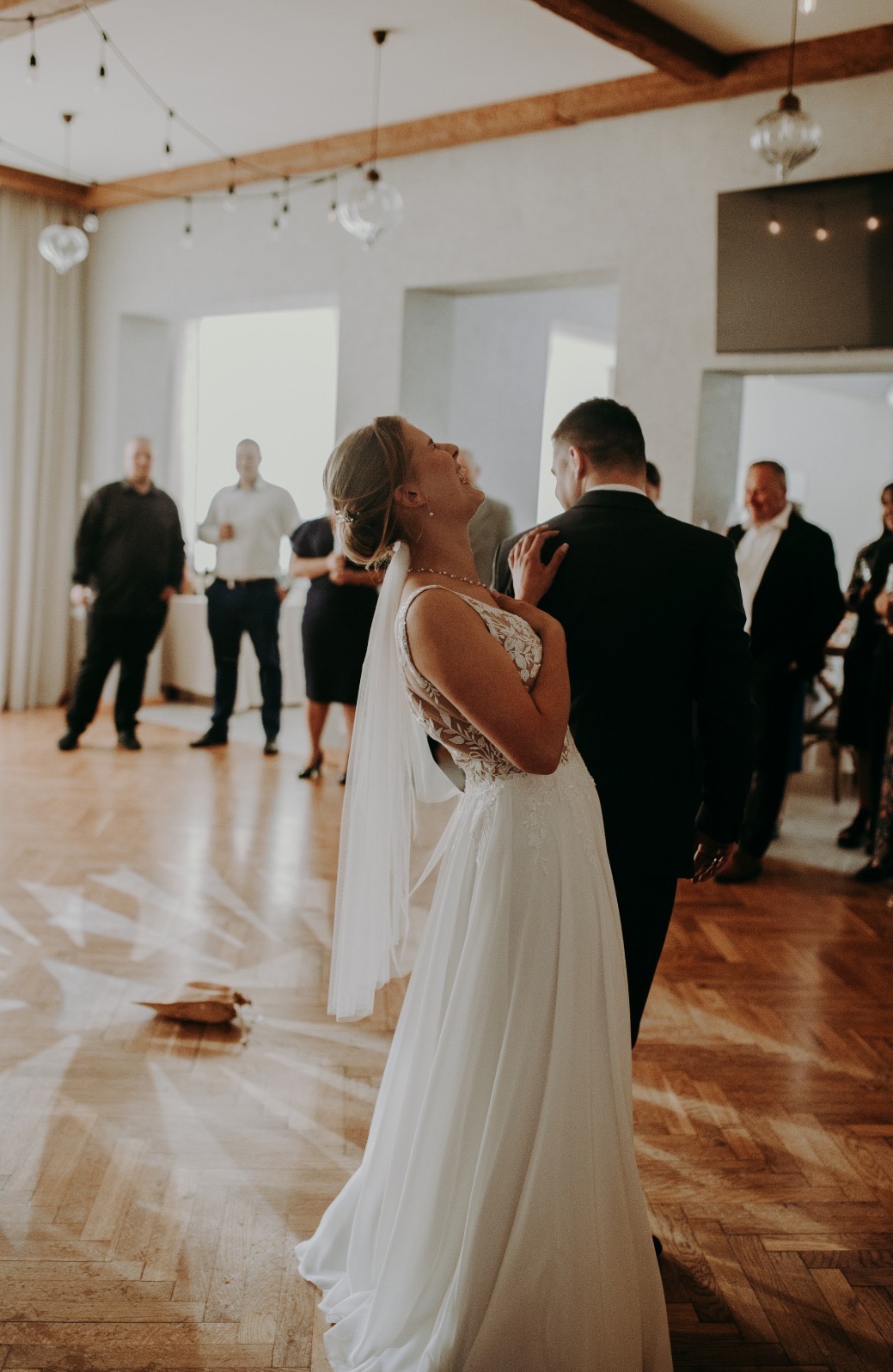 Uśmiechnięta panna młoda w białej sukni tanecznej, z welonem, opiera się o ramię pana młodego podczas wesela. W tle goście i drewniana podłoga.