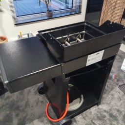 Pan Wkręcik - Czarny grill gazowy na kółkach, z rozłożonym bocznym blatem, widoczny w pomieszczeniu, z pomarańczowym przewodem gazowym i elementami montażowymi na podłodze.