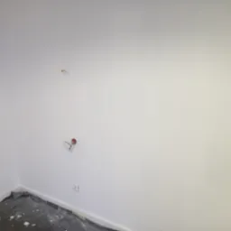 Świeżo pomalowane na biało wnętrze z wystającymi instalacjami i zabezpieczoną podłogą. Widok na narożnik pokoju z gładką, jednolitą powierzchnią ścian.