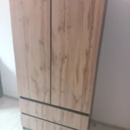 Zamontowana, dwudrzwiowa szafa z trzema szufladami w kolorze drewna, z ciemnymi obramowaniami, na tle białej ściany i jasnej podłogi.