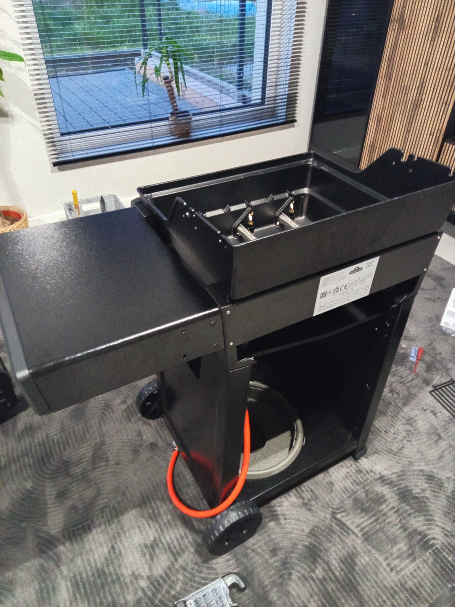 Czarny grill gazowy na kółkach, z rozłożonym bocznym blatem, widoczny w pomieszczeniu, z pomarańczowym przewodem gazowym i elementami montażowymi na podłodze.