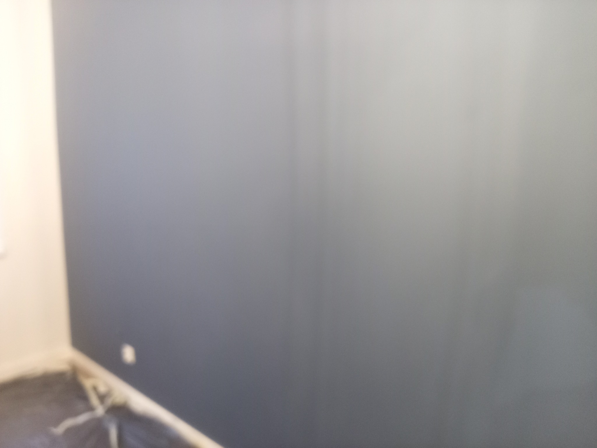 Ściana pomalowana na szaro, z widocznym gniazdkiem elektrycznym i zabezpieczoną folią podłogą. Nierównomierne oświetlenie.