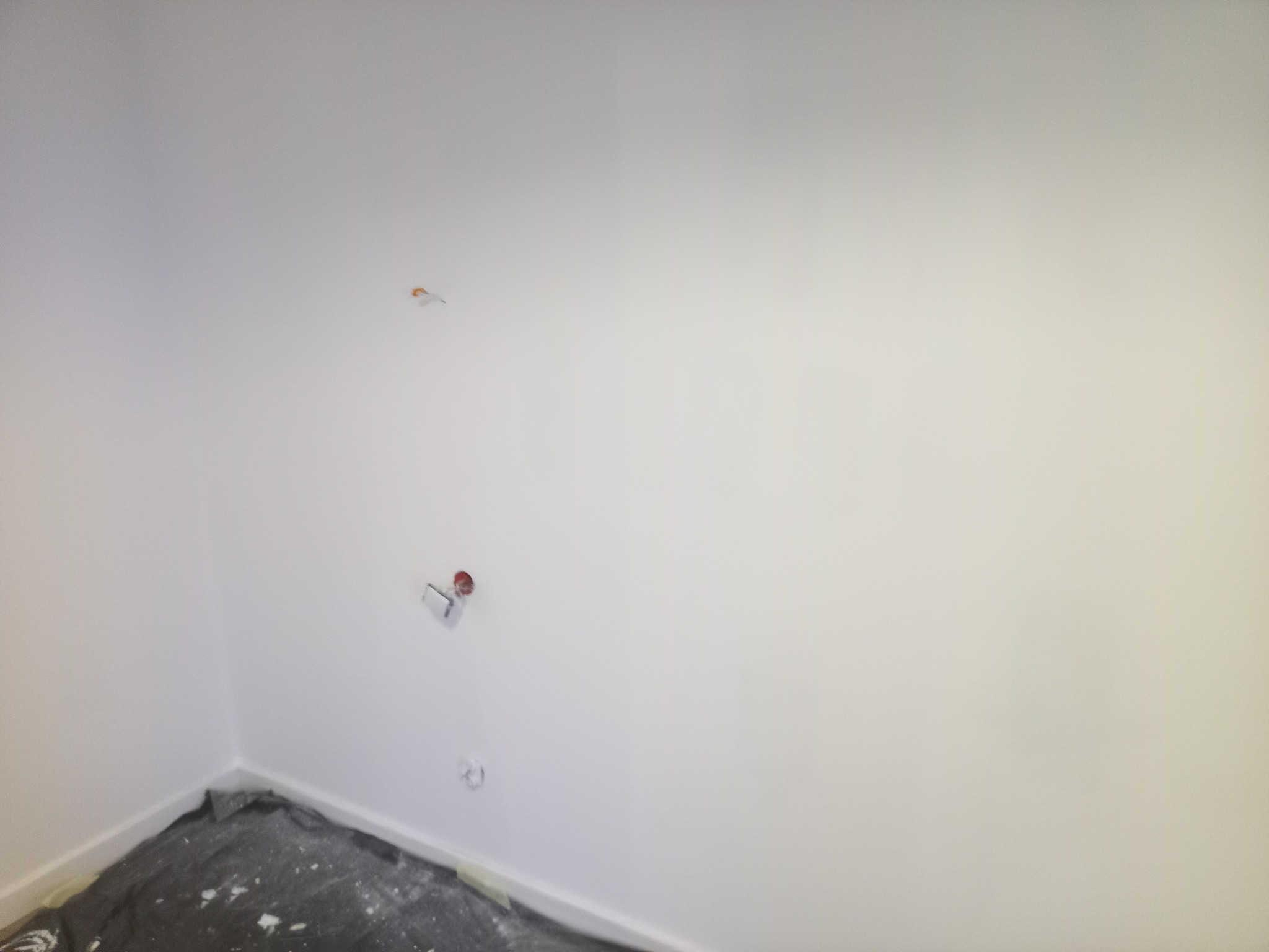 Świeżo pomalowane na biało wnętrze z wystającymi instalacjami i zabezpieczoną podłogą. Widok na narożnik pokoju z gładką, jednolitą powierzchnią ścian.