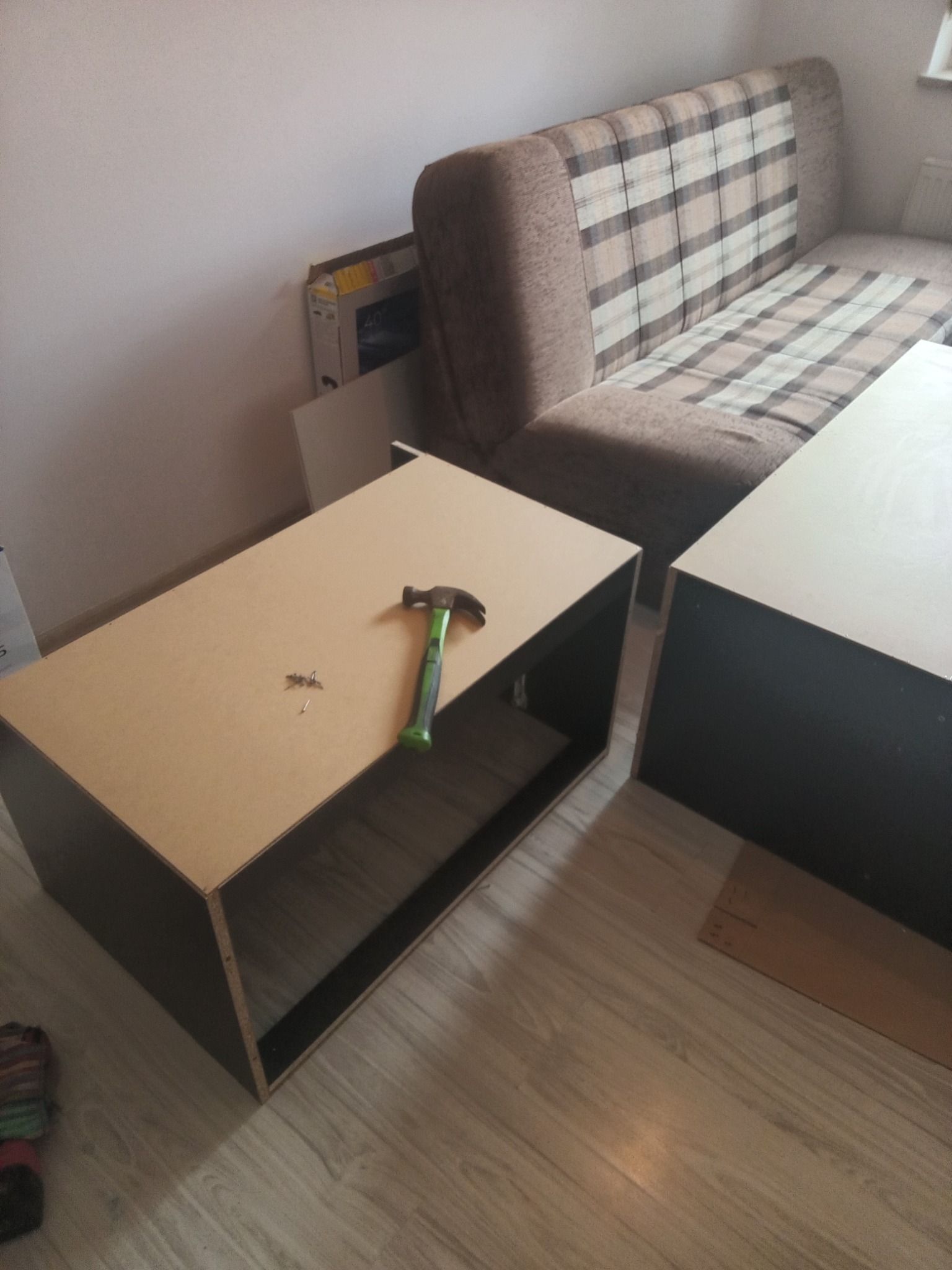 Składanie szafki: młotek i gwoździe na blacie. Widoczne elementy meblowe i sofa w tle. Etap montażu, surowe wykończenie, wnętrze mieszkania.