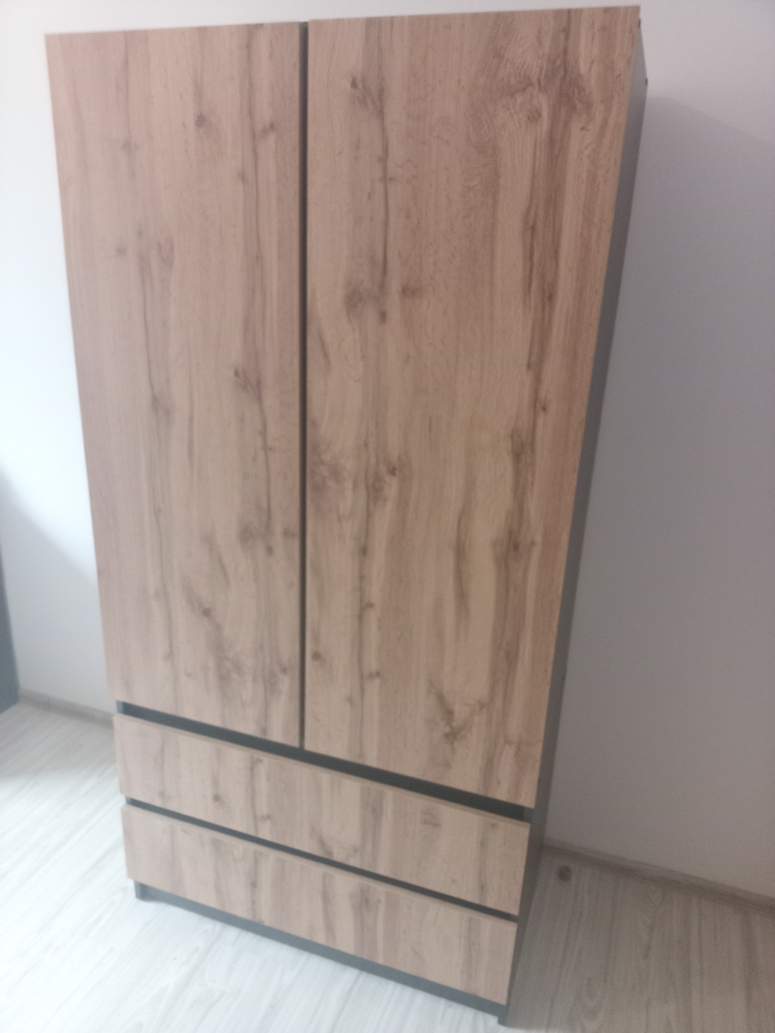 Zamontowana, dwudrzwiowa szafa z trzema szufladami w kolorze drewna, z ciemnymi obramowaniami, na tle białej ściany i jasnej podłogi.