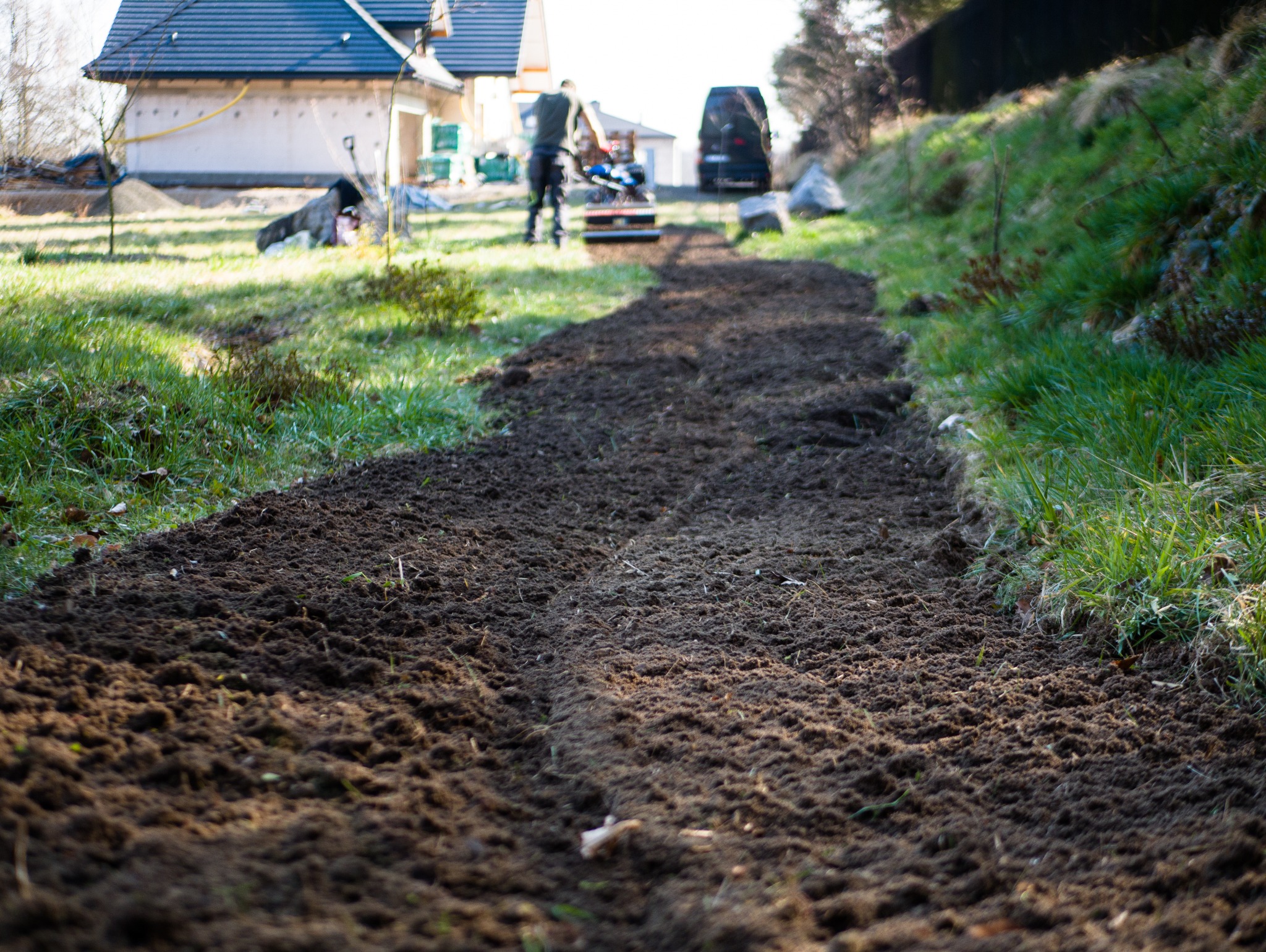 Przygotowanie gruntu pod trawnik: wyrównana ziemia po użyciu glebogryzarki, widoczna granica z istniejącą trawą, w tle dom i pracownik z maszyną.