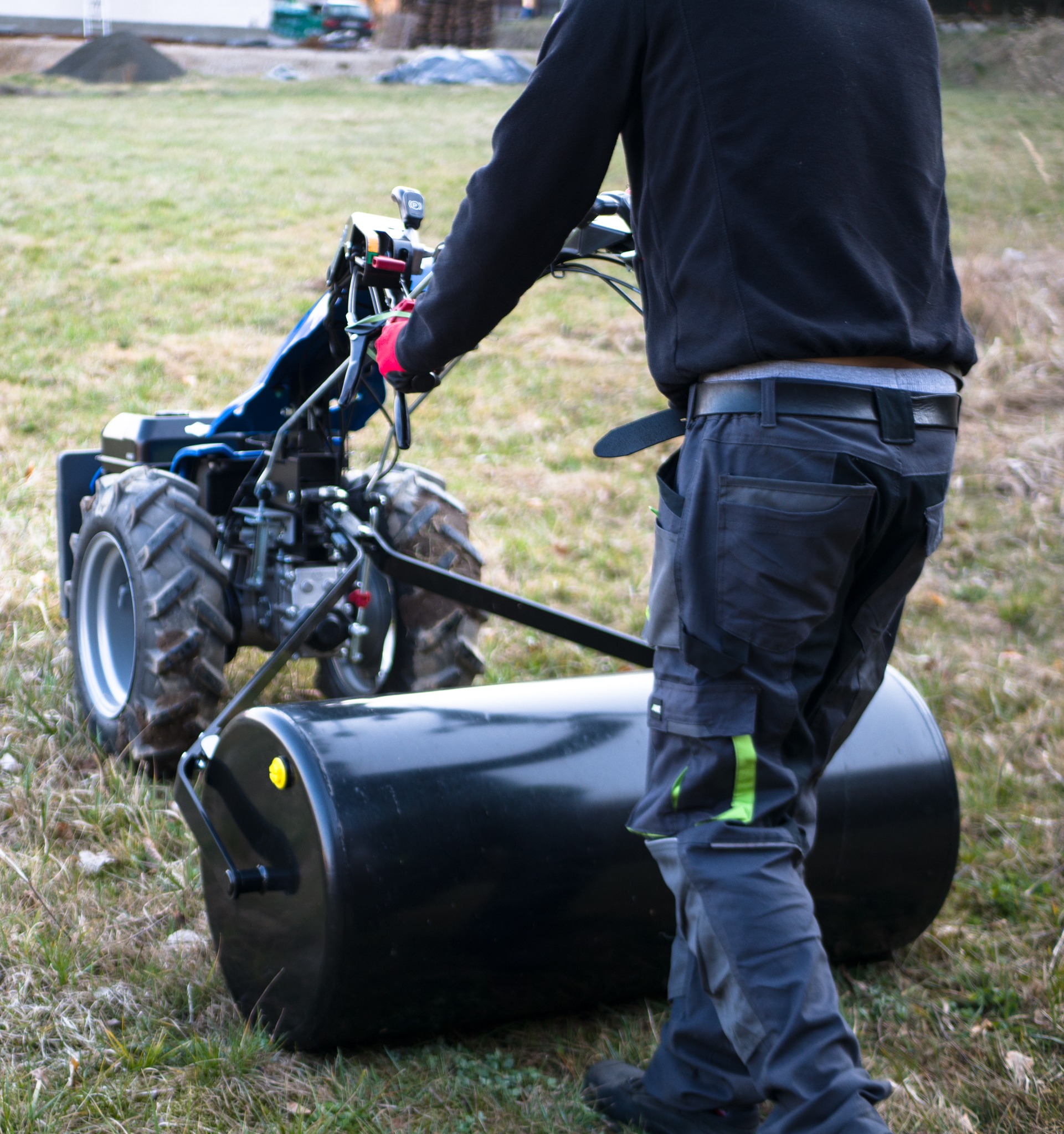 Pracownik w odzieży roboczej obsługuje traktor z wałem, wyrównując teren zielony. W tle widoczny kopiec ziemi i zieleń.