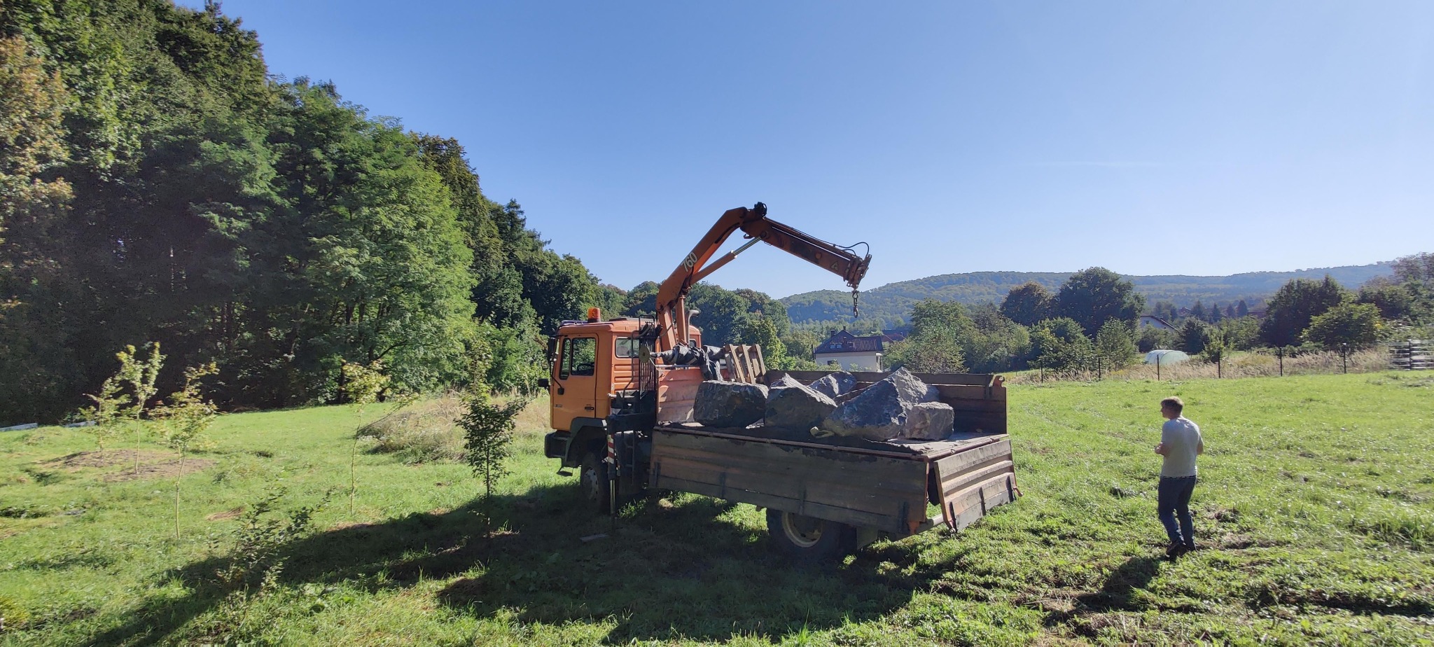 Pomarańczowy dźwig samochodowy z ładunkiem dużych kamieni na łące, w tle zalesione wzgórza i osoba w oddali. Słoneczny dzień, widoczne drzewa i trawa.