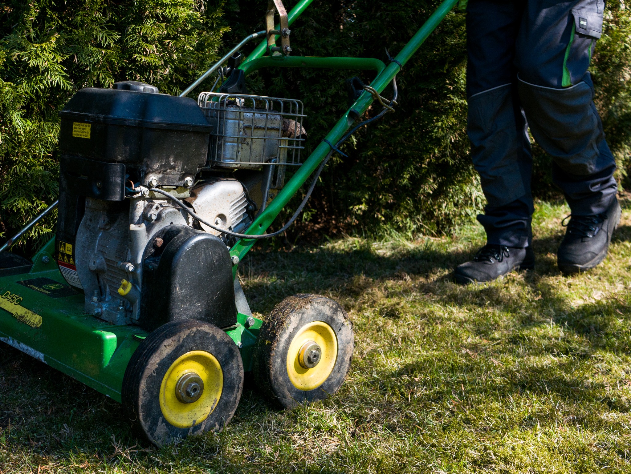 Zielona kosiarka spalinowa z żółtymi kołami na trawniku, częściowo widoczne nogi operatora w roboczych spodniach i butach, krzew w tle.