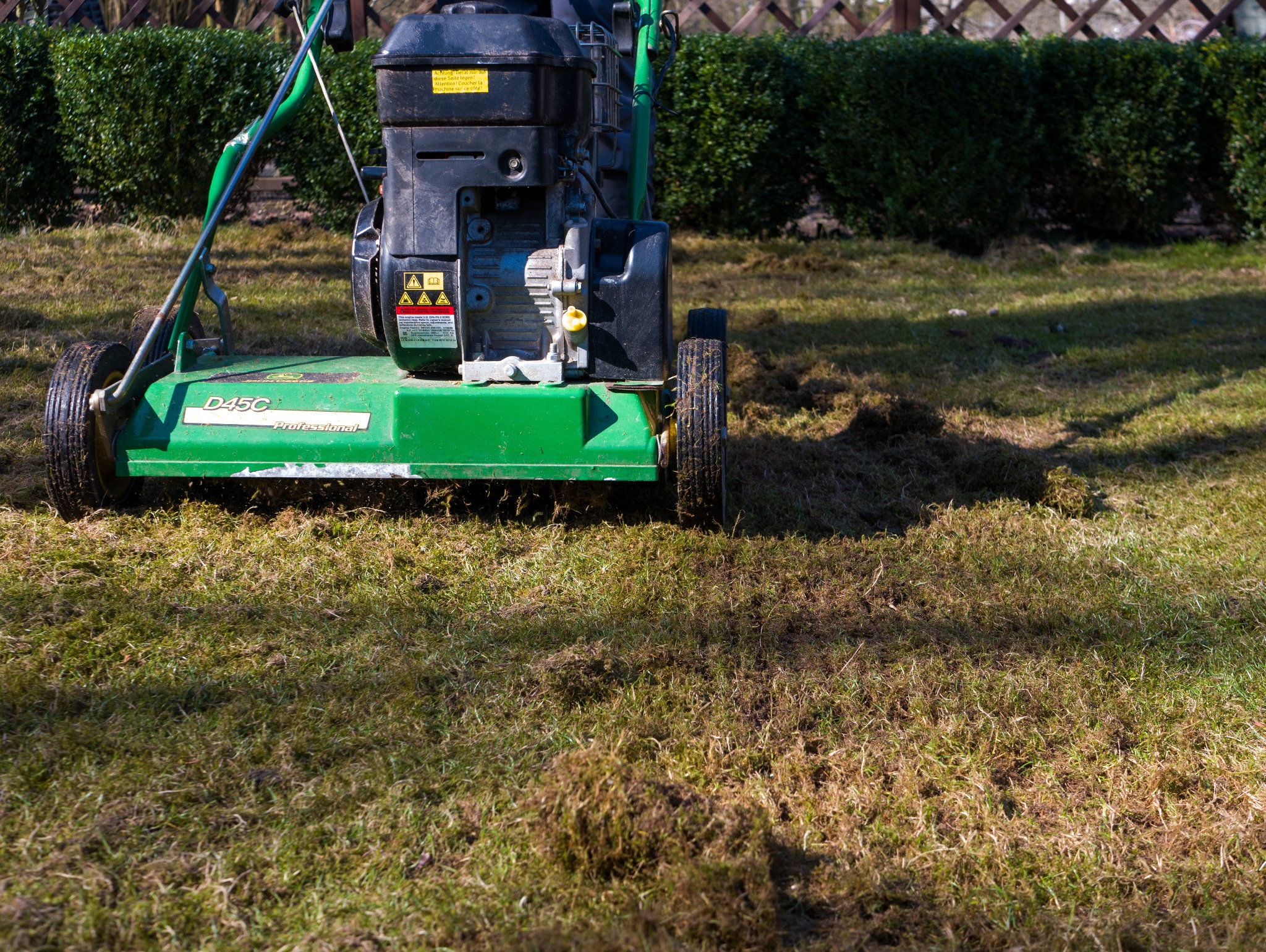 Profesjonalna kosiarka D45C na trawniku po wertykulacji, widoczne resztki mchu i darni. W tle żywopłot i cień drzew. Słoneczny dzień.