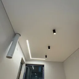 Biały sufit z oświetleniem LED w kształcie litery L i trzema punktowymi lampami. Widoczny fragment instalacji wentylacyjnej i okno w tle. Minimalistyczny design.