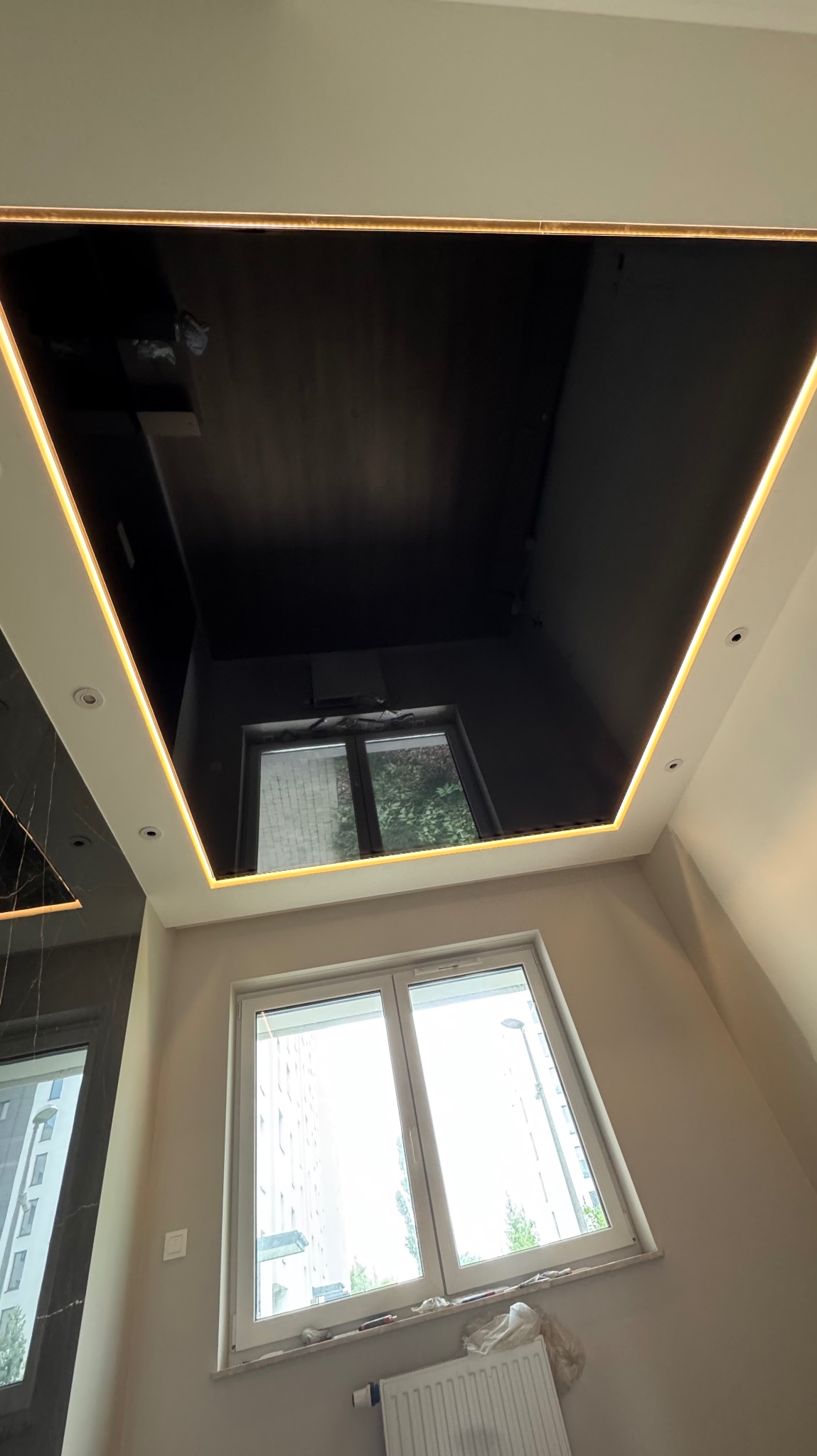 Czarny, błyszczący sufit z oświetleniem LED, odbijający widok okna. Widok z dołu na nowoczesne wykończenie wnętrza, z widocznym grzejnikiem i oknem w tle.