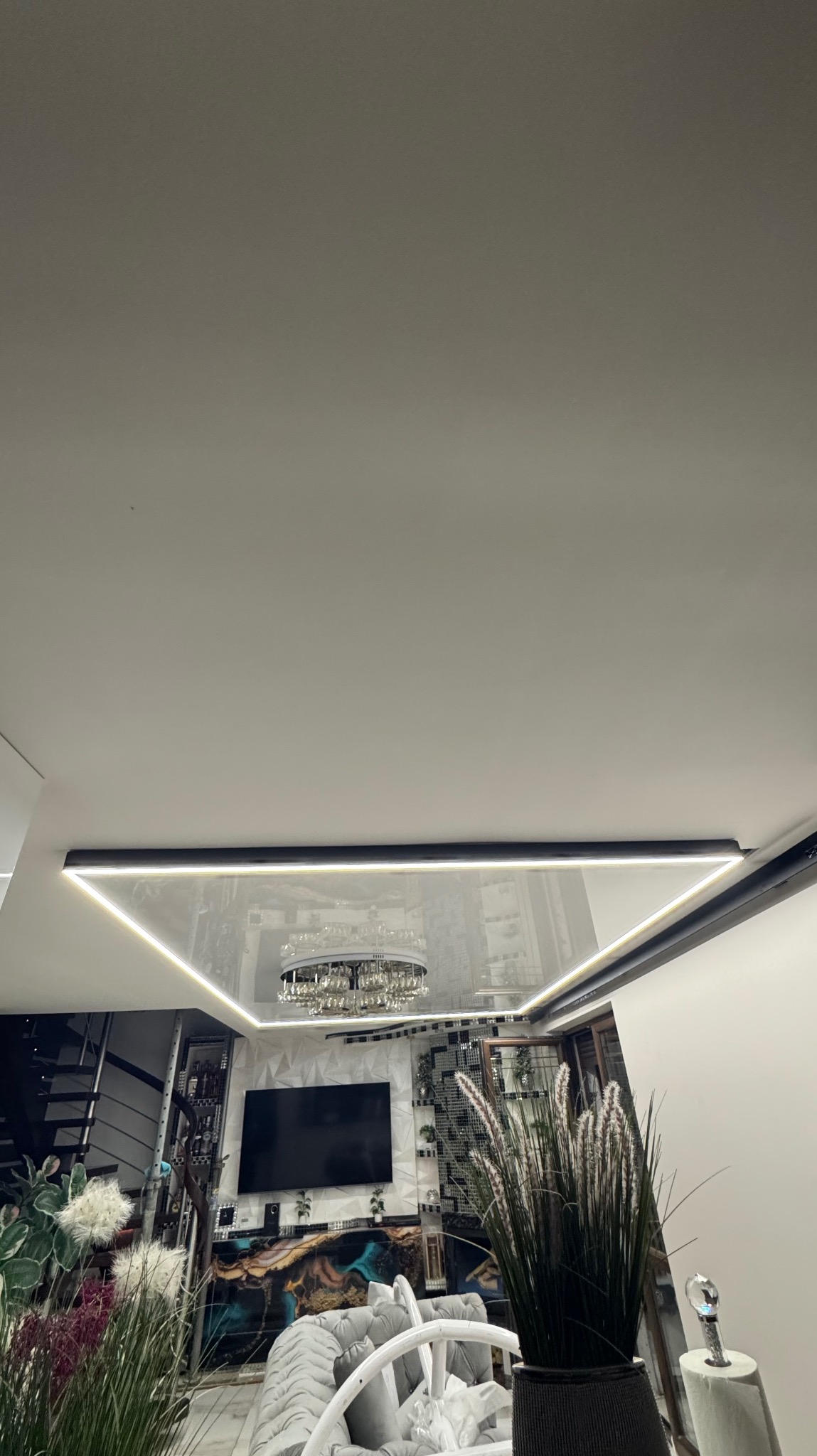 Nowoczesny salon z podświetlanym, trójkątnym sufitem z efektem lustra, w którym odbija się kryształowy żyrandol i elementy dekoracyjne wnętrza.